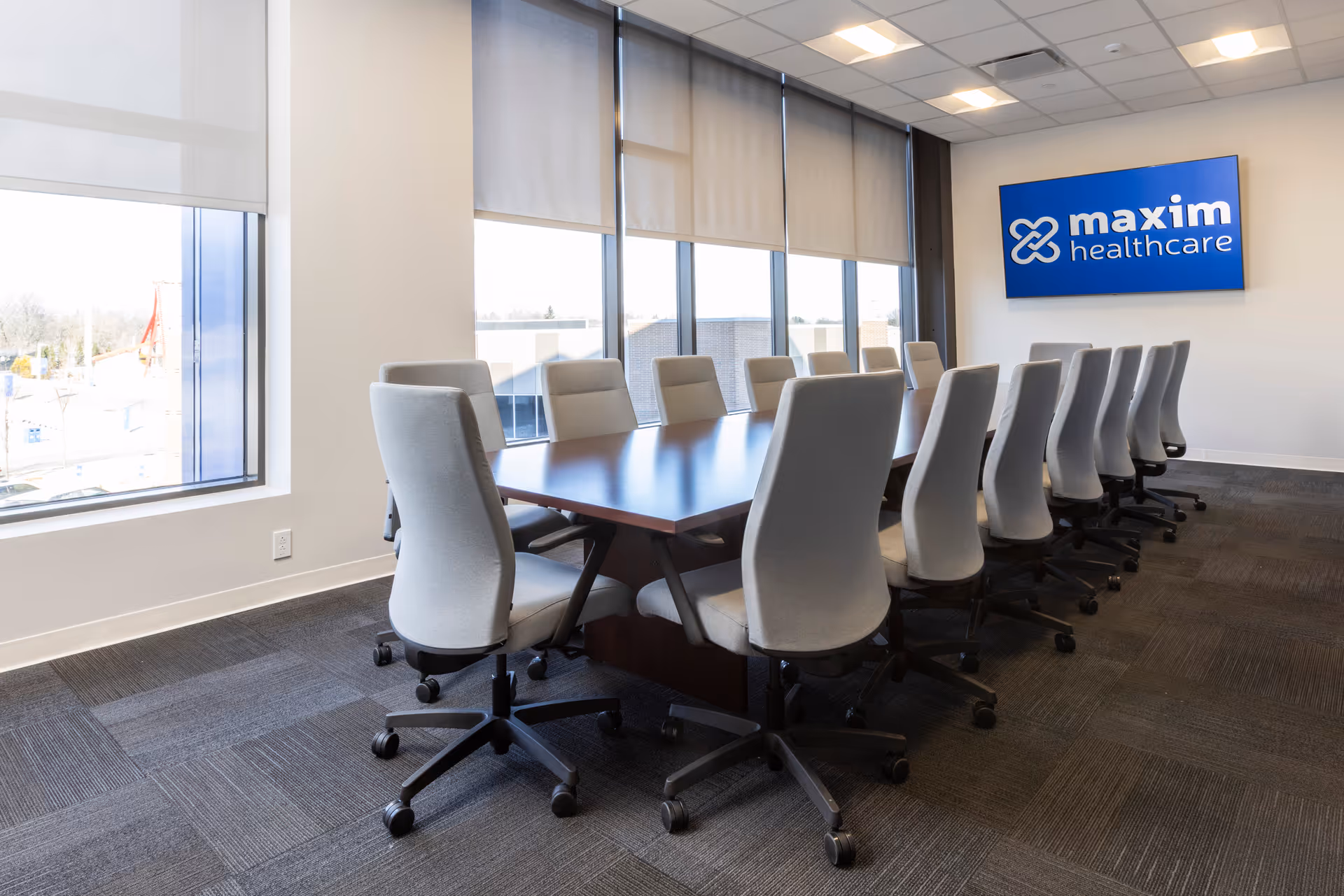 Large conference room with long table, gray chairs, windows, and Maxim Healthcare sign.