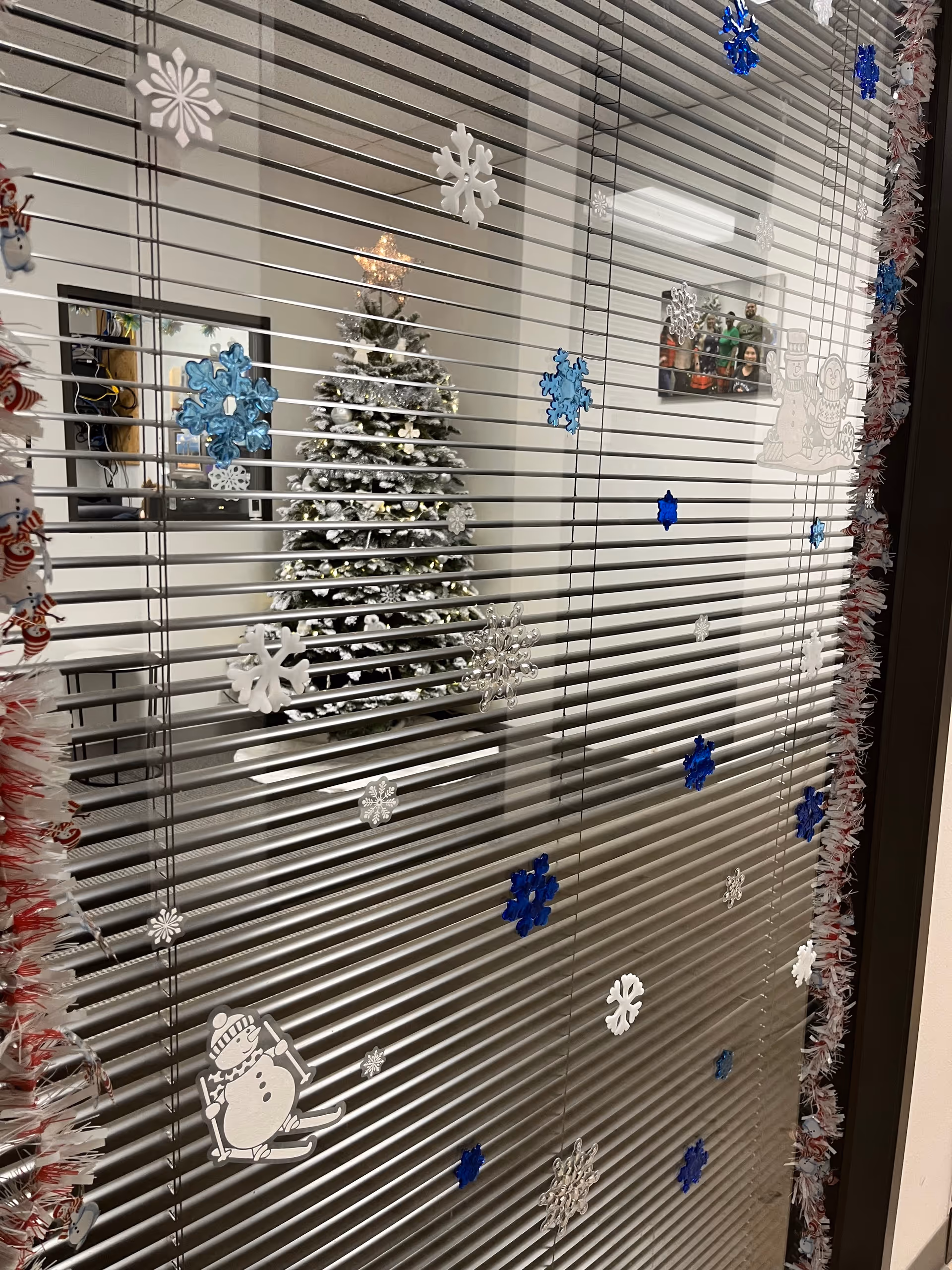 Window blinds decorated with snowflakes reveal a Christmas tree inside.