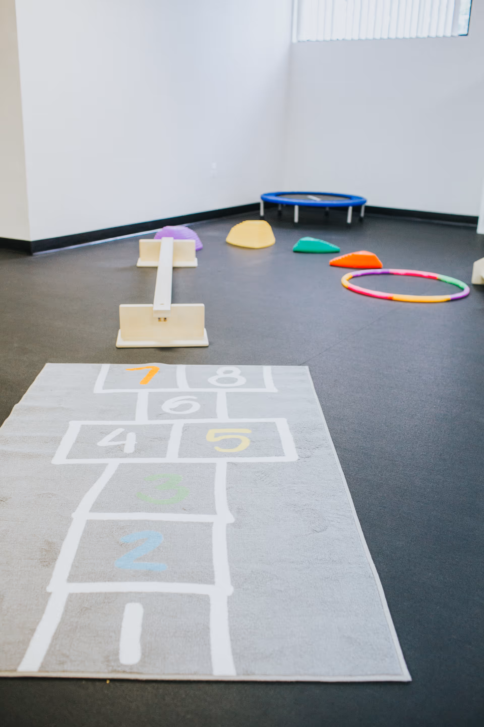 Indoor therapy room with hopscotch rug, balance beam, trampoline, and colorful foam shapes.