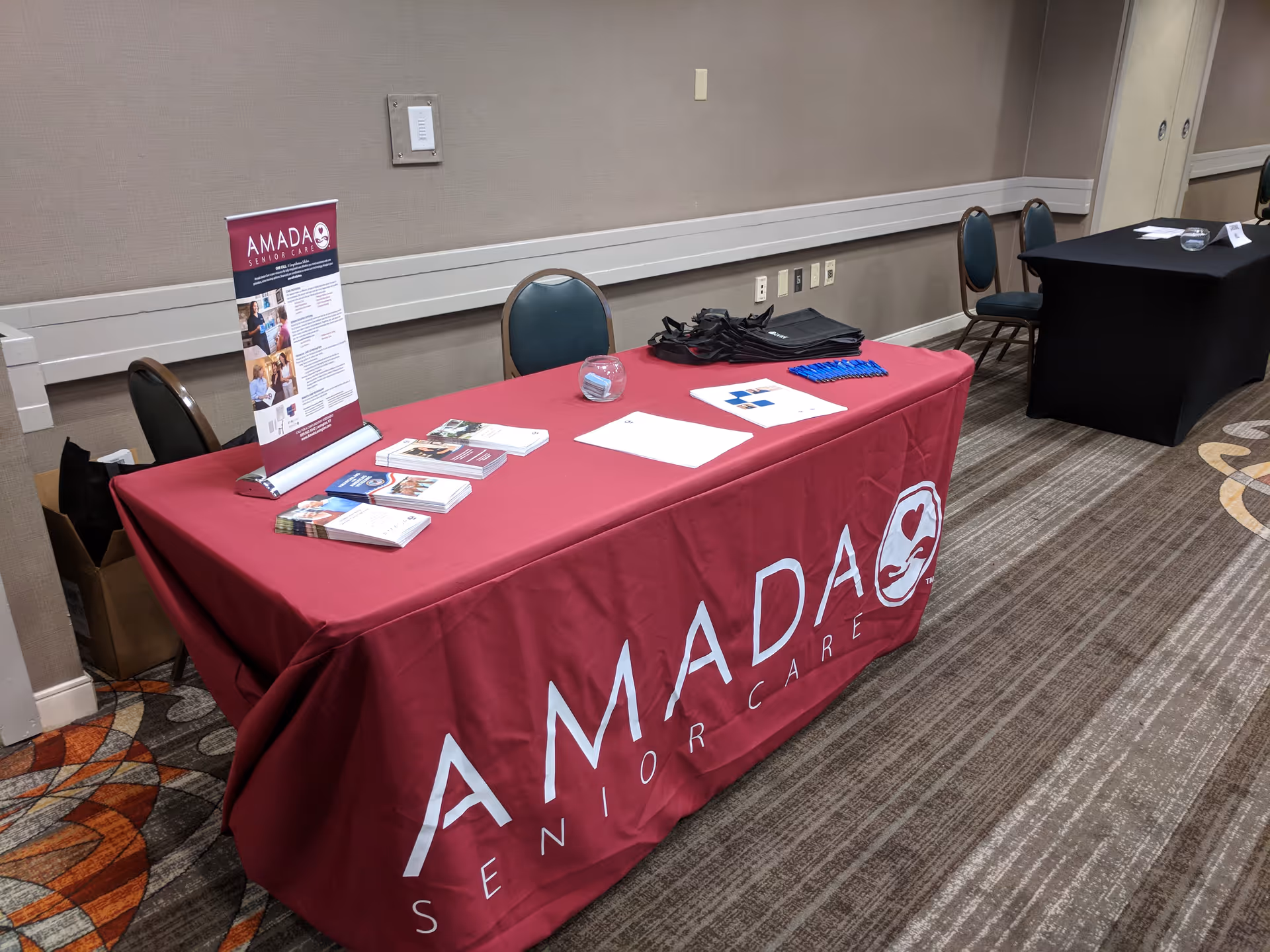 An Amada Senior Care booth with a red tablecloth and brochures in an interior conference area.