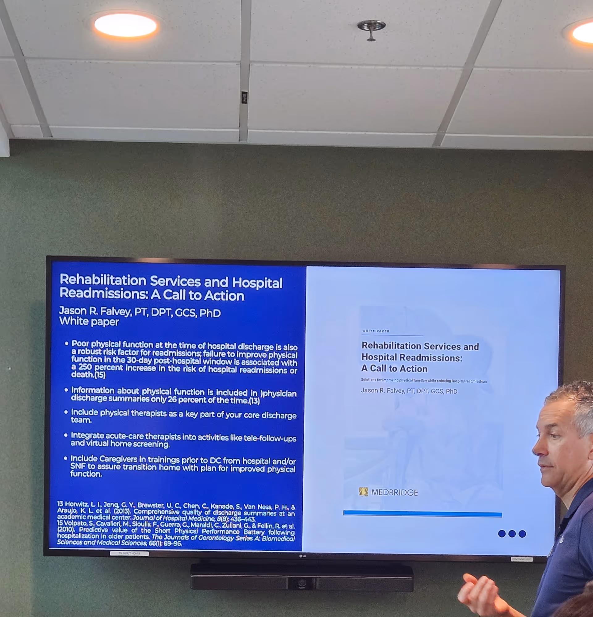 A large wall-mounted screen displays a blue presentation slide as a man speaks indoors.
