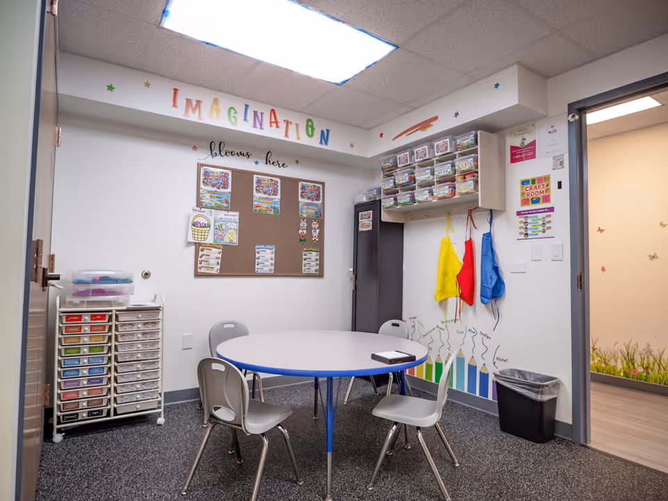 Bright craft room with a round table, small chairs, storage bins, and wall art.