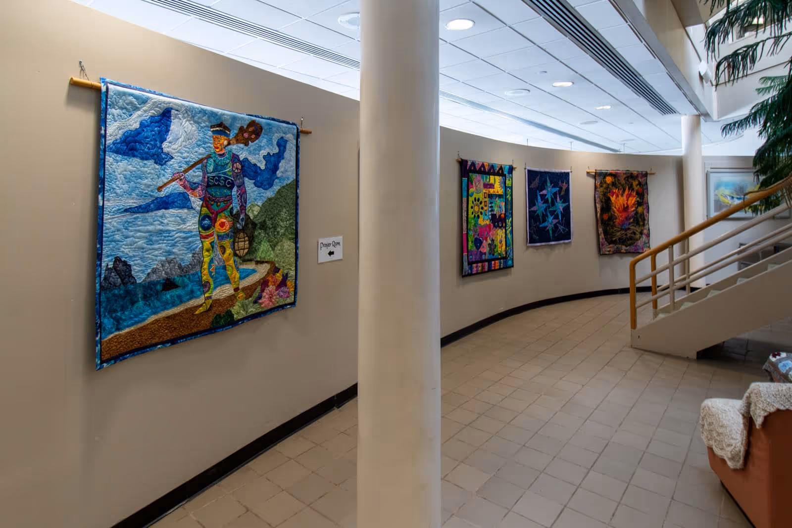 Long curved hospice hallway with colorful wall quilts and a staircase.