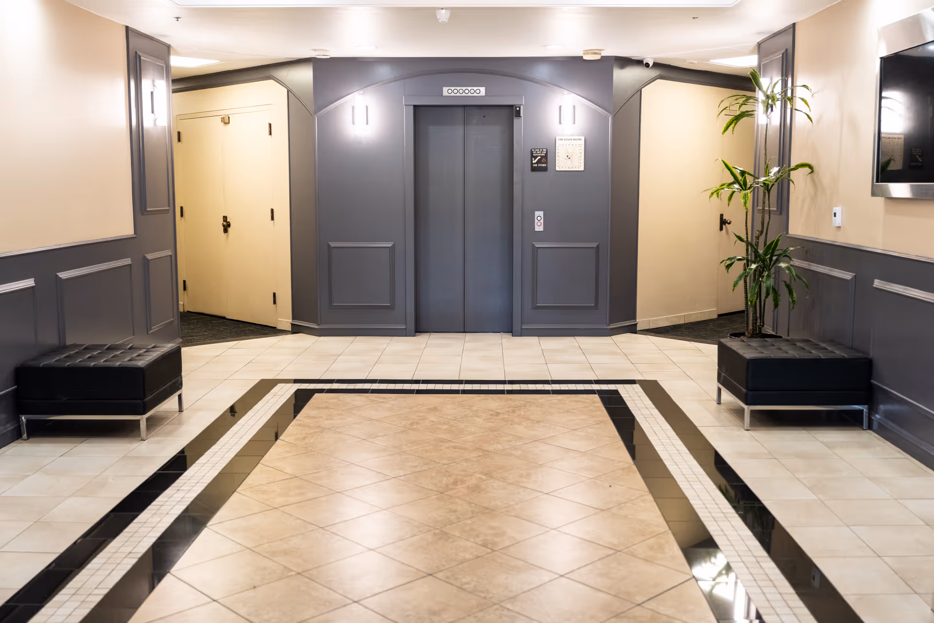 Lobby hallway with central elevator, beige walls, tiled floor, black benches, and a plant.