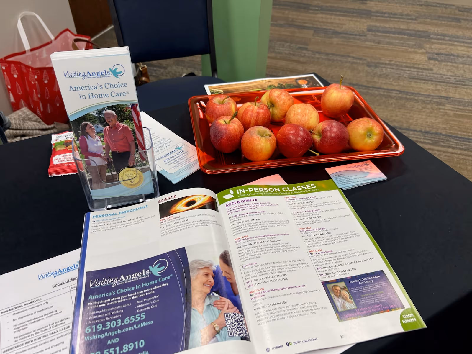 Indoor table with Visiting Angels brochures, a red apple tray, and promotional materials.