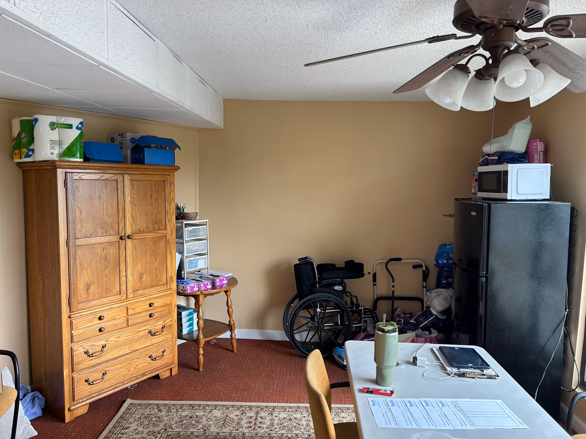 A cluttered senior living room with a wooden cabinet, wheelchair, refrigerator, microwave, and table.