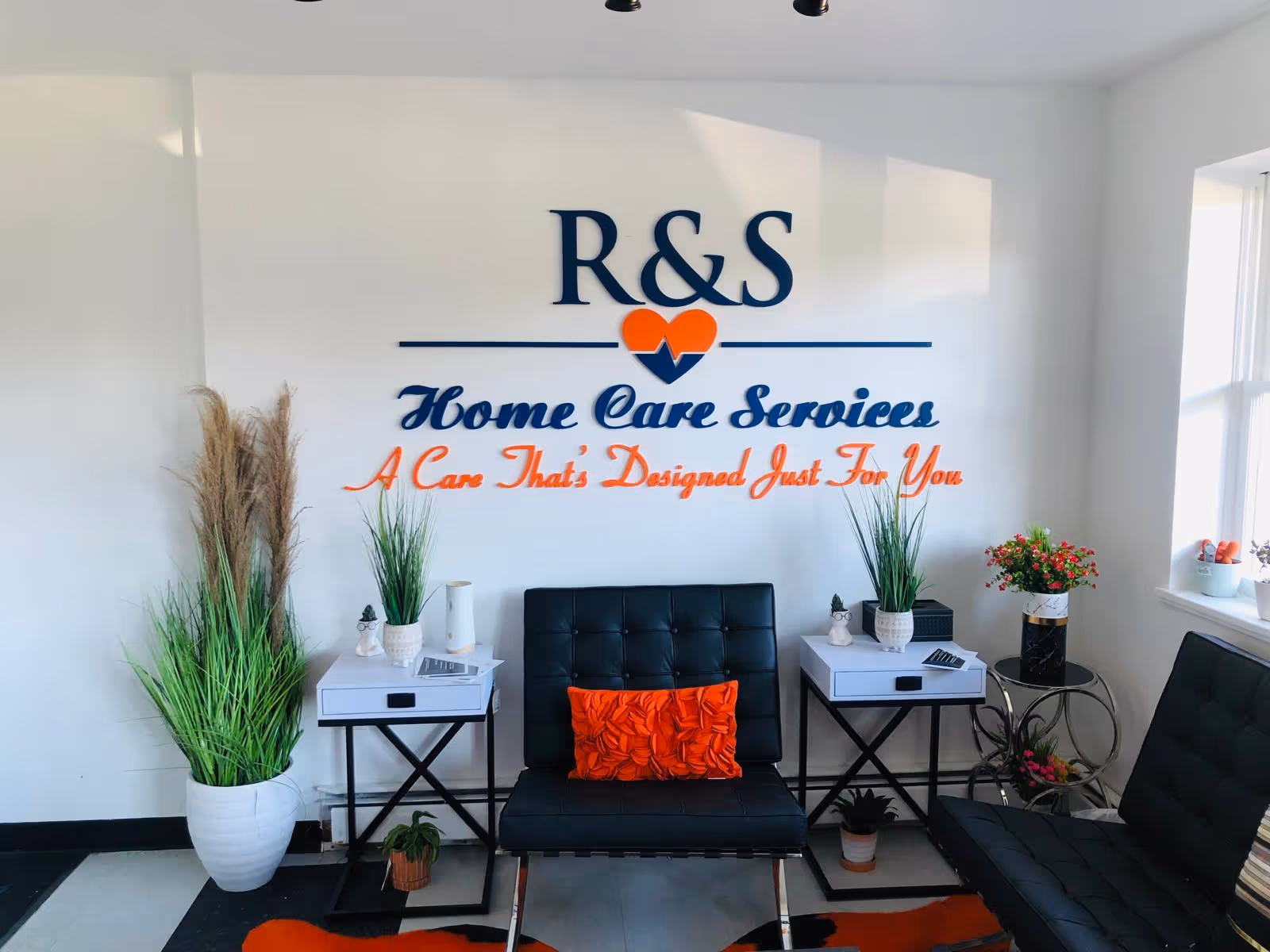 Lobby area with R&S Home Care Services sign, black tufted couch, white tables, and plants.