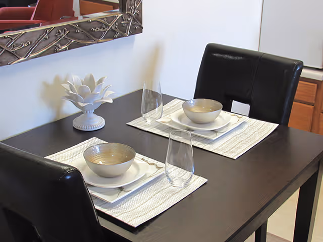Two-place dining table set with bowls, glasses, and a decorative plant near a mirror.