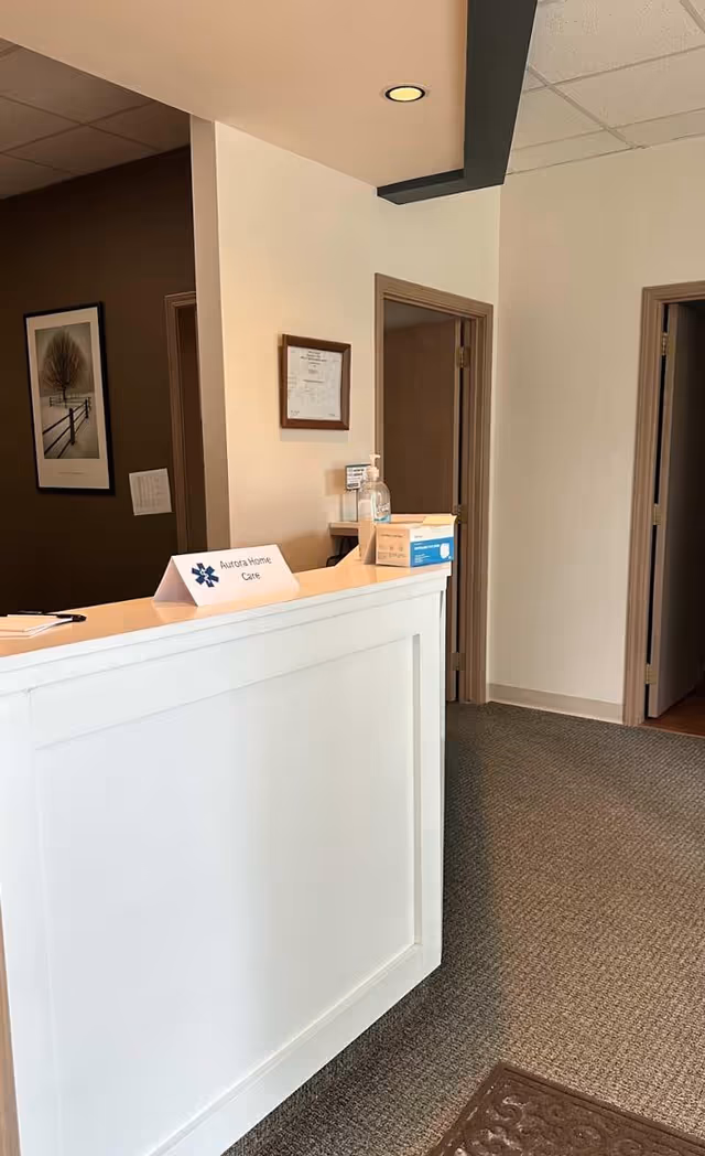 Reception desk in a neutral lobby with an Aurora Home Care sign and sanitizer.