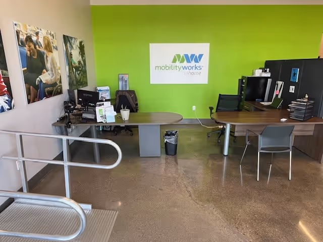 A bright green wall with the MobilityWorks logo behind two desks and office equipment.