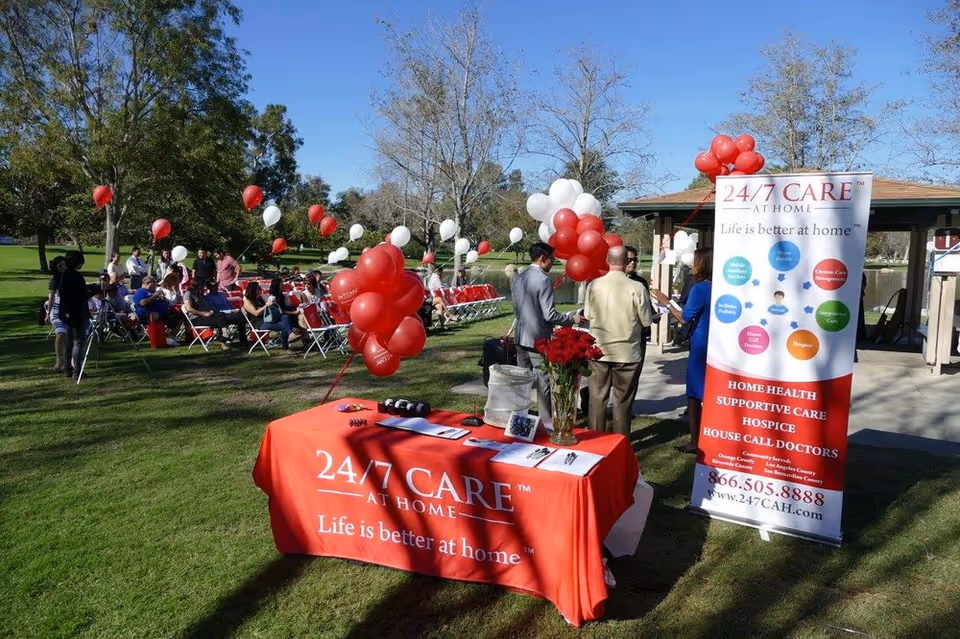Outdoor event at 24/7 Care At Home with a red table, balloons, and attendees.