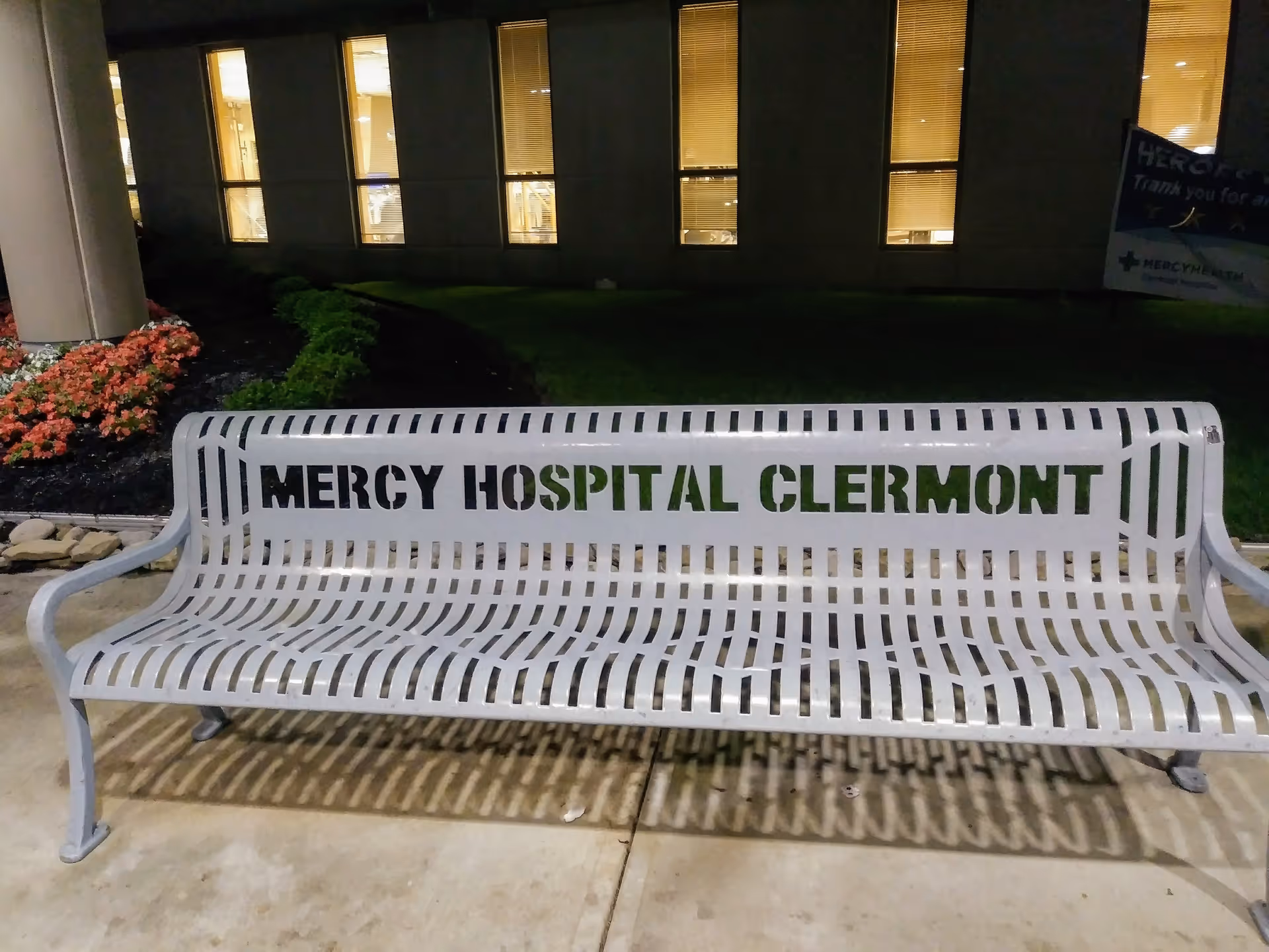 Nighttime view of a white metal bench with Mercy Hospital Clermont signage outside.