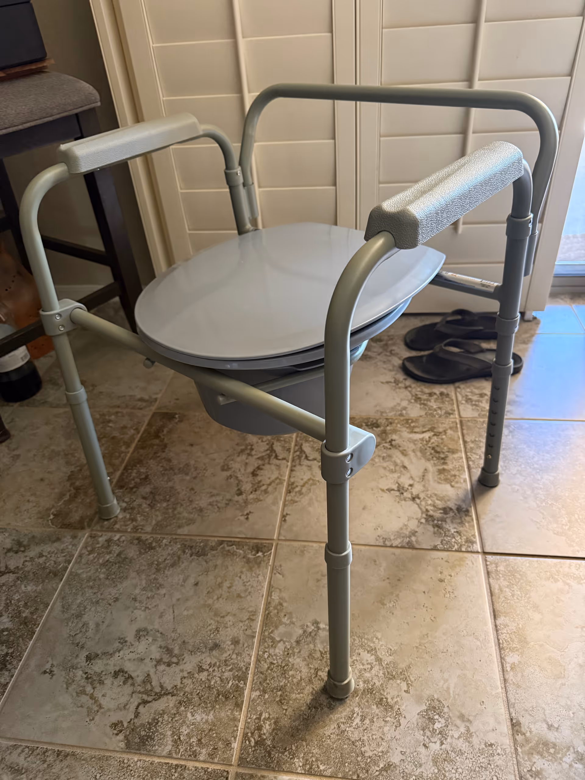 Light gray commode chair with armrests in a tiled bathroom.