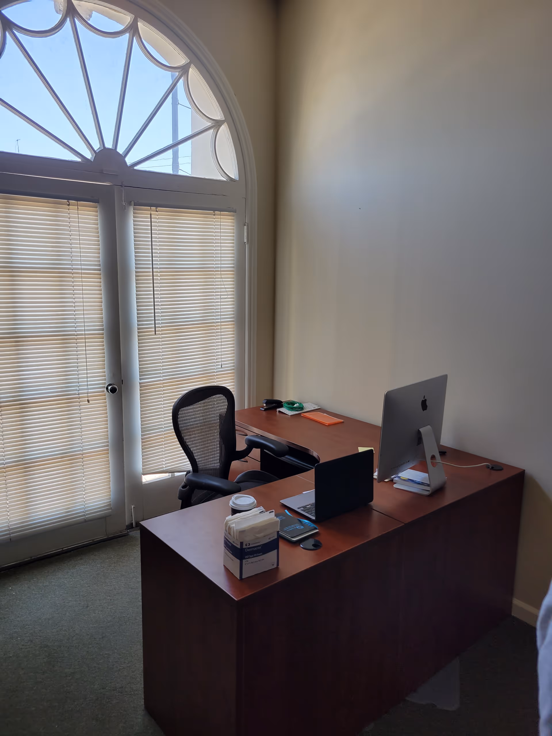 Office desk with computer setup beside a large arched window with blinds.