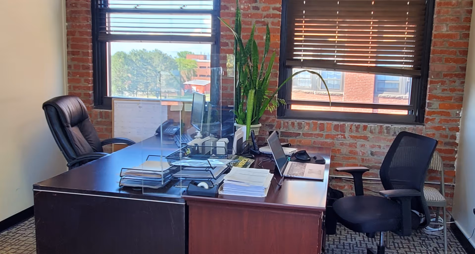 Office workspace with dark wood desk, brick walls, blinds, a laptop, and a plant.