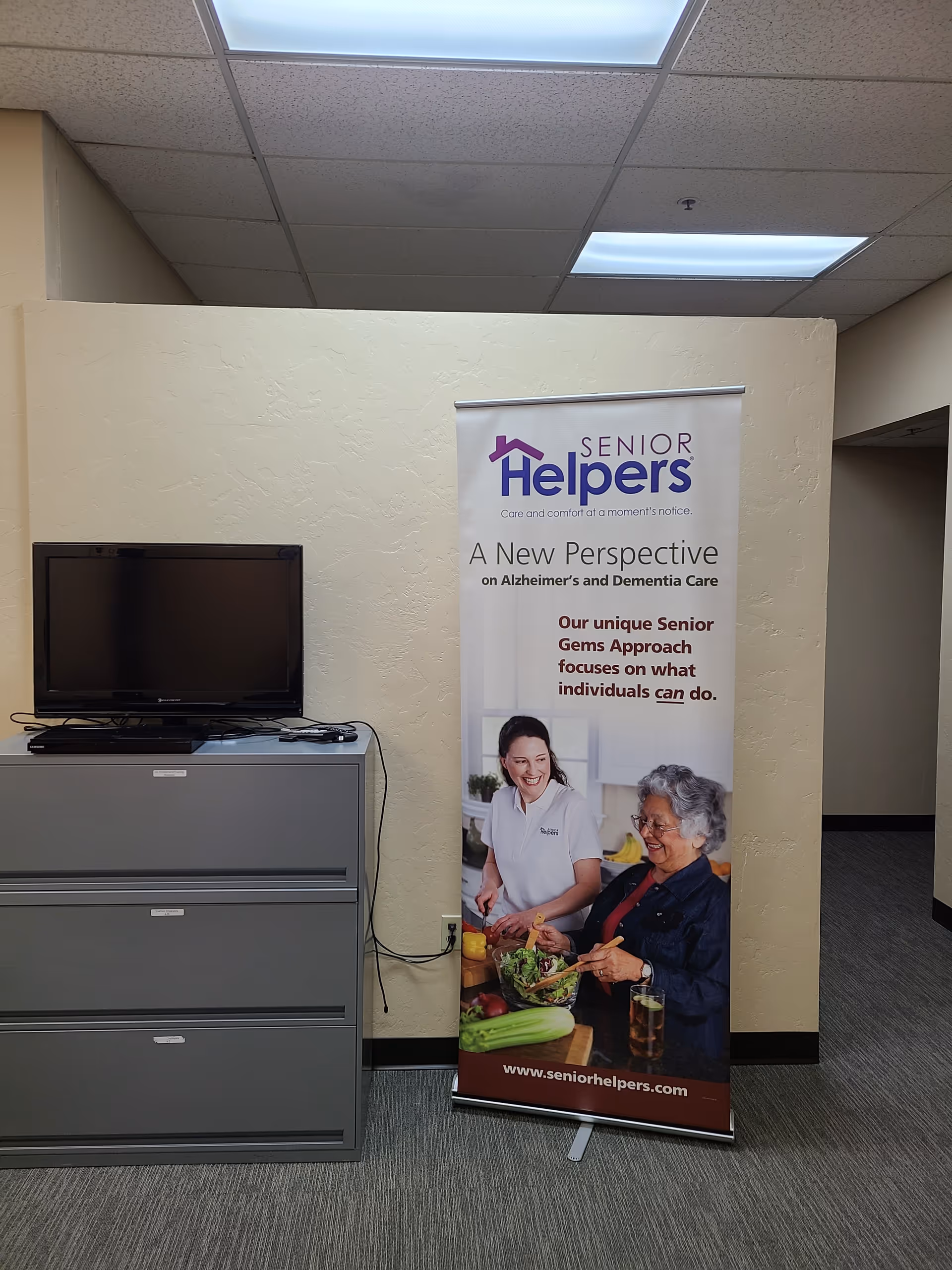 Indoor office area with a Senior Helpers banner beside a filing cabinet and television.