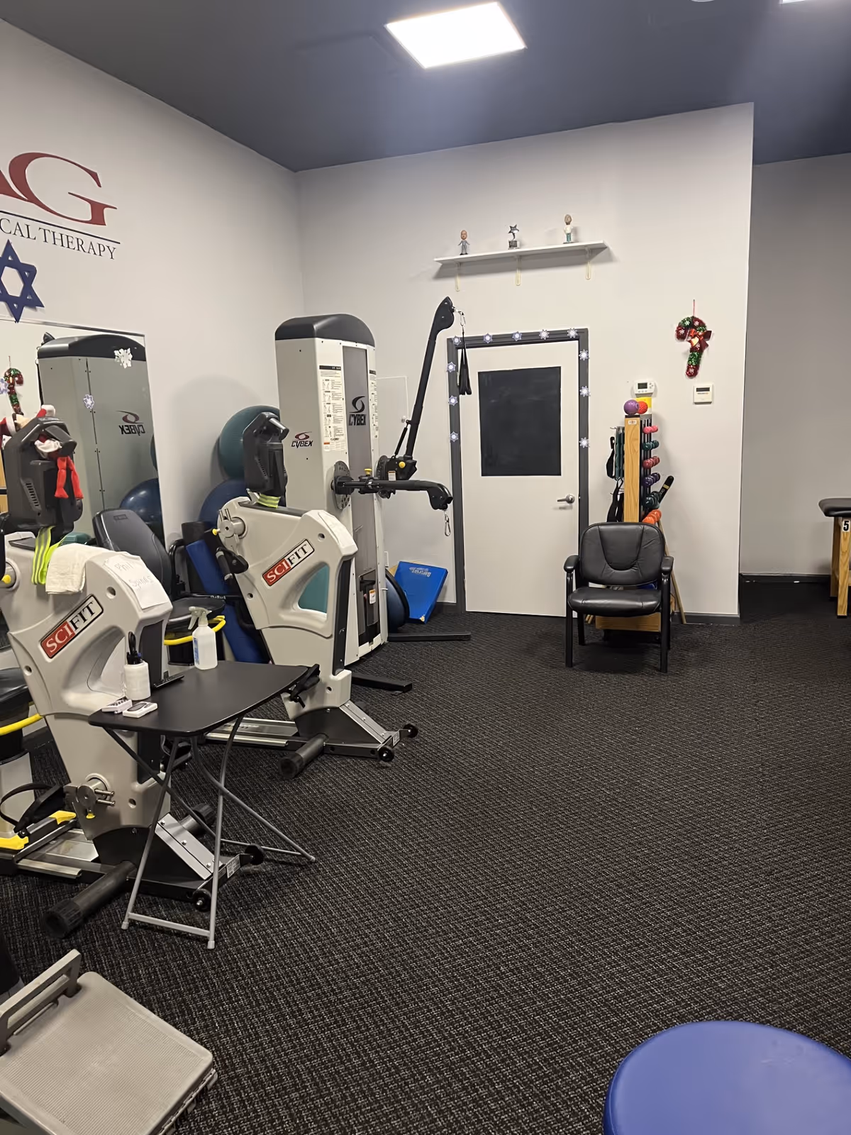 Interior physical therapy gym with SCIFIT machines, exercise equipment, and a chair.