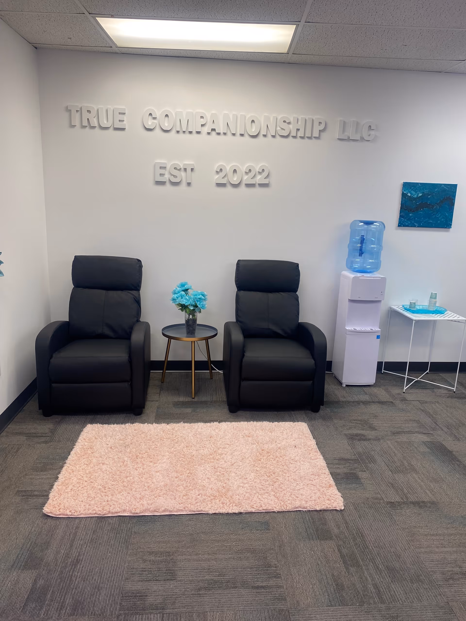 Lobby seating area with two black recliners, a small table with blue flowers, water cooler.
