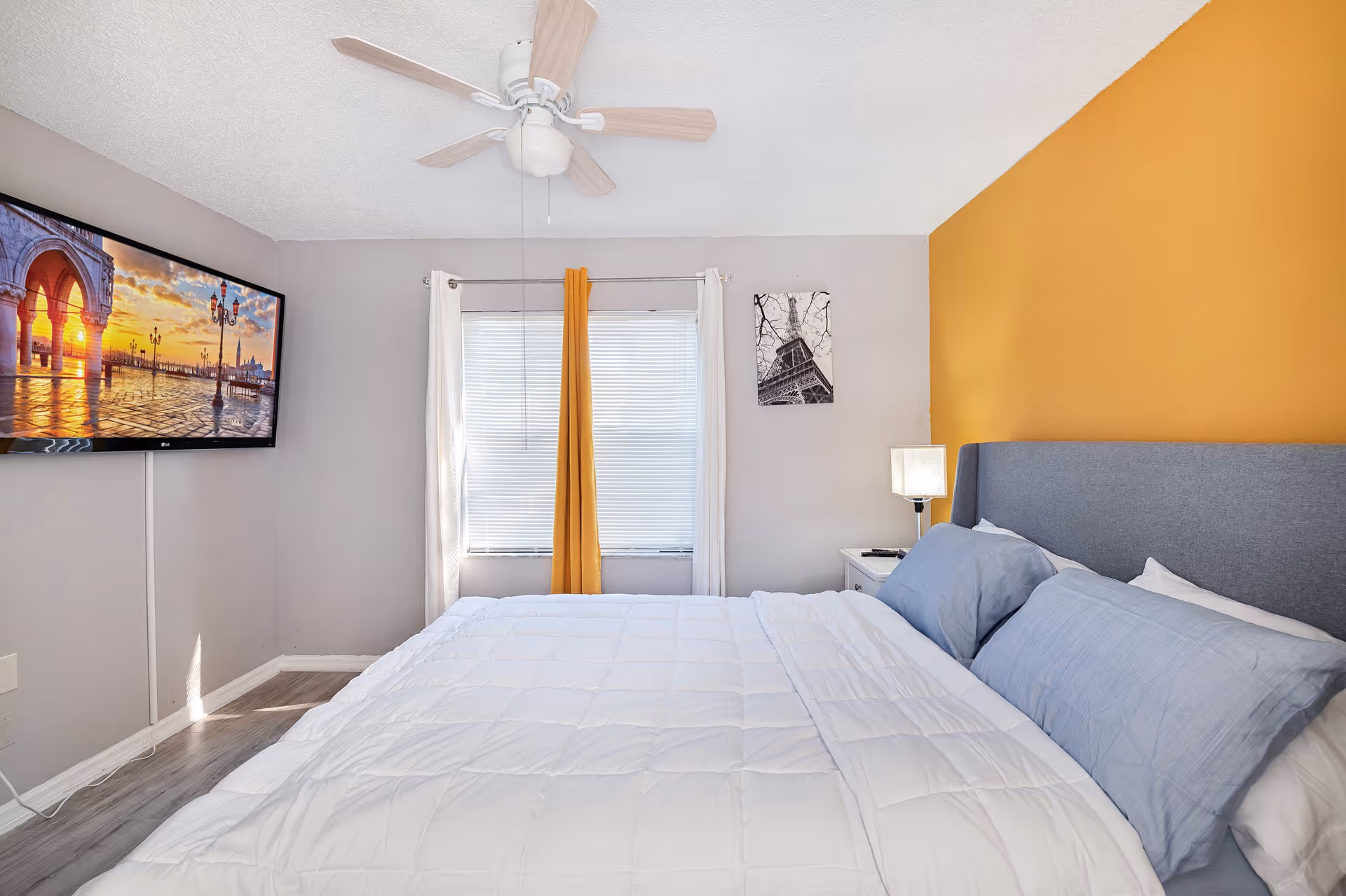 Bright bedroom with a large bed, yellow accent wall, ceiling fan, and mounted TV.