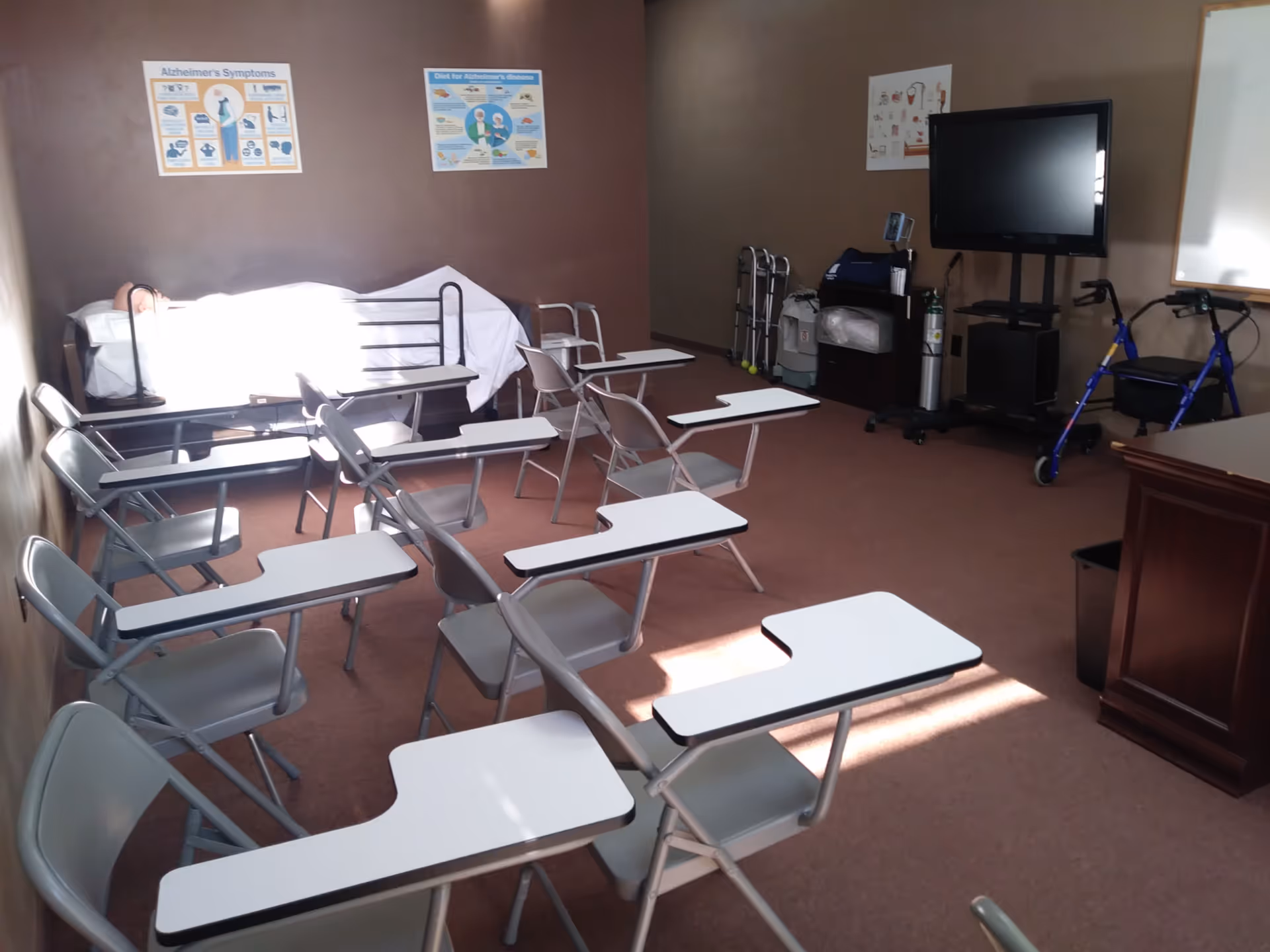 Interior classroom-like room with fold-out desks, chairs, a bed, medical equipment, and a TV.