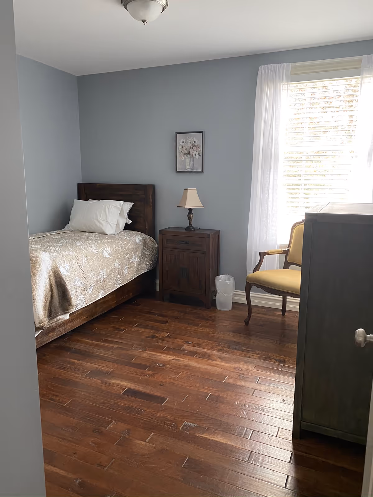 A tidy bedroom in a senior living facility with wood floors, bed, nightstand, lamp, and window.