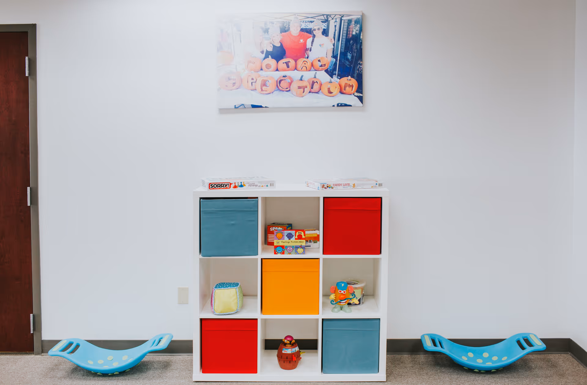 A white cubby shelf with colorful fabric bins and toys against a plain wall.