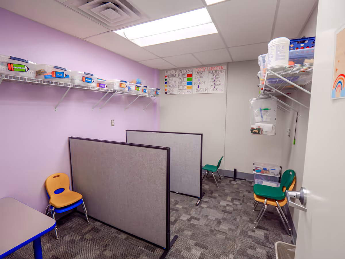 Purple-walled activity room with cubicle partitions, shelves of bins, and chairs.