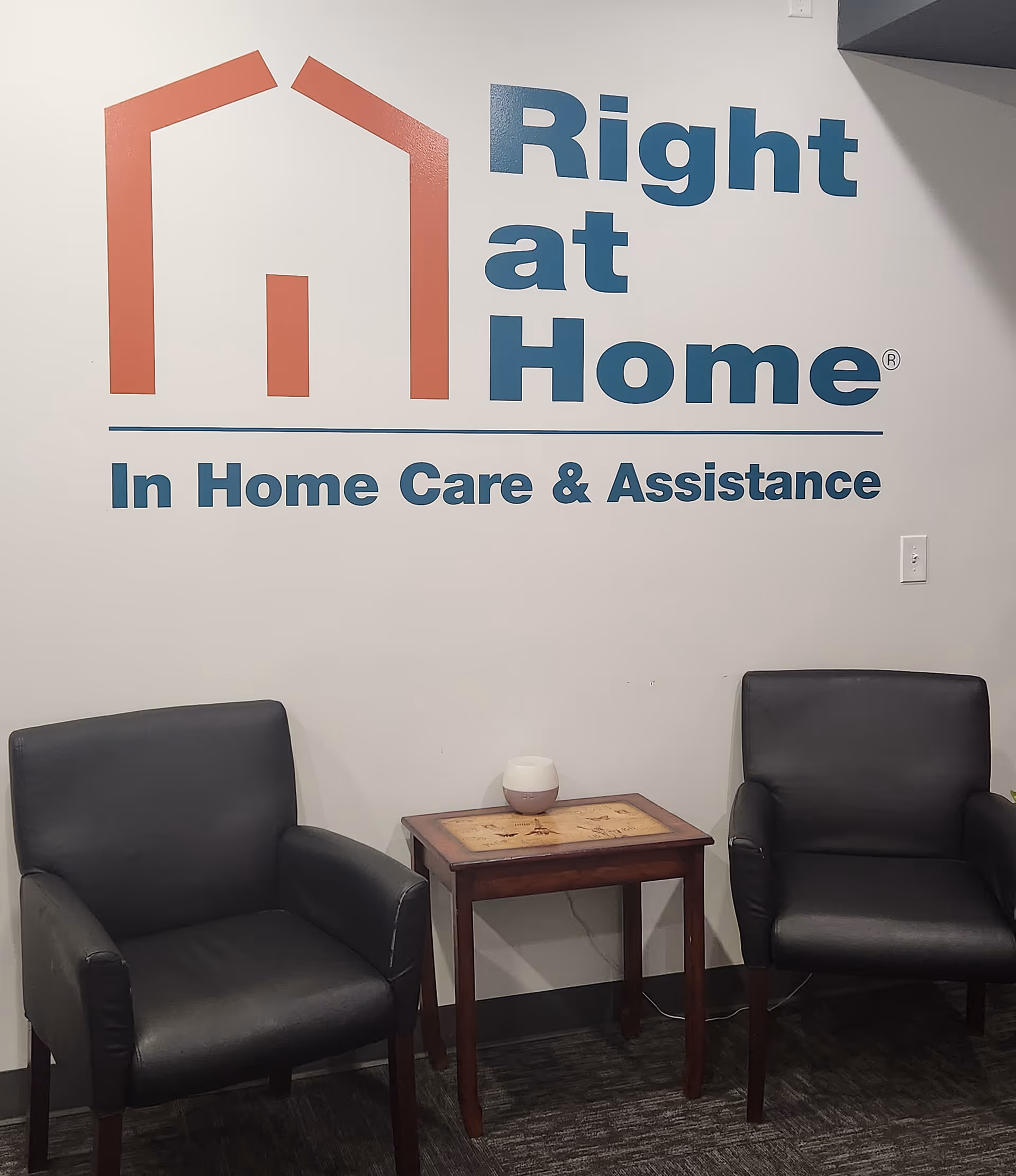 Two black chairs flank a wooden table in front of a Right at Home sign.