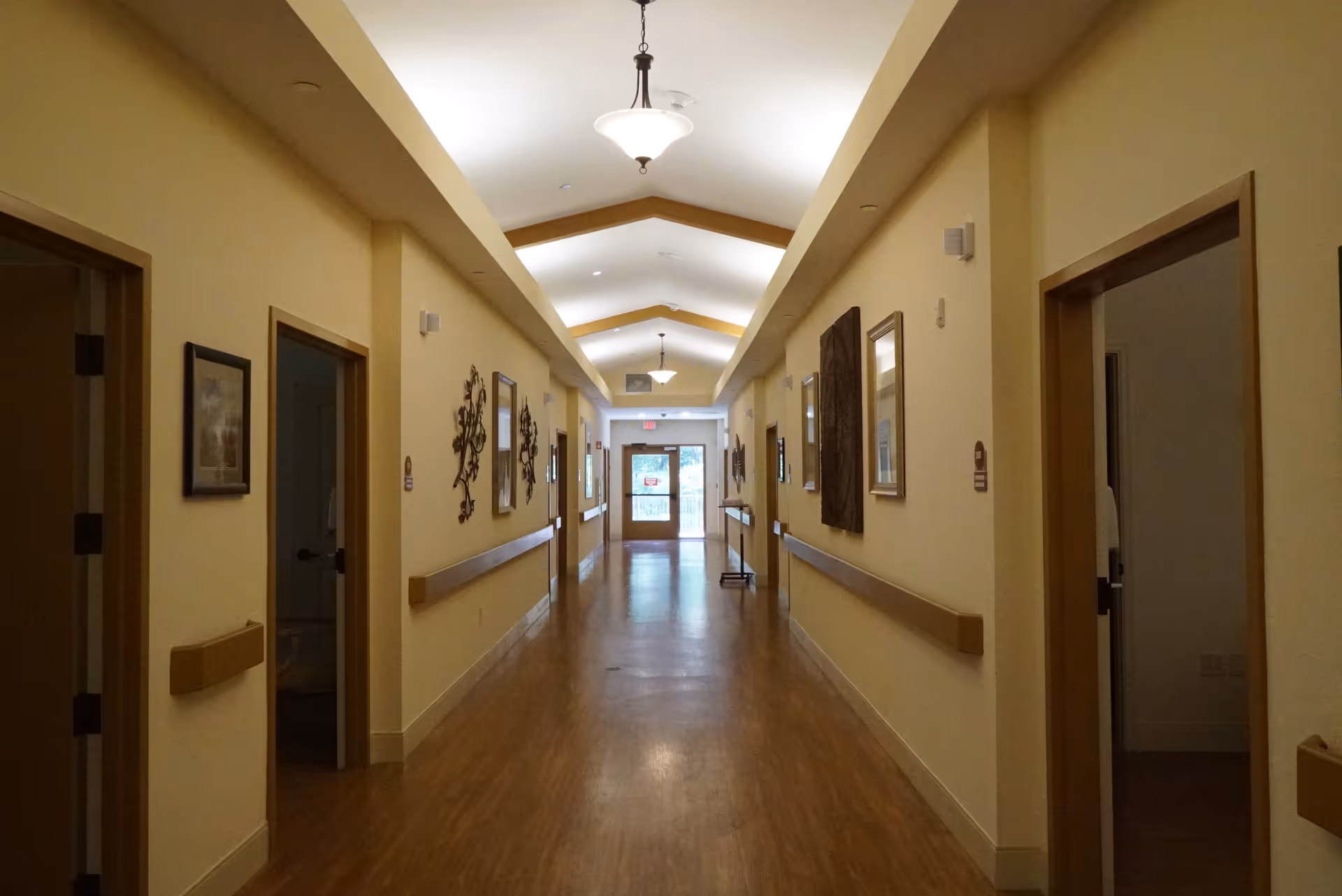 Long, well-lit hallway in a senior living facility with doors, railings, and artwork.