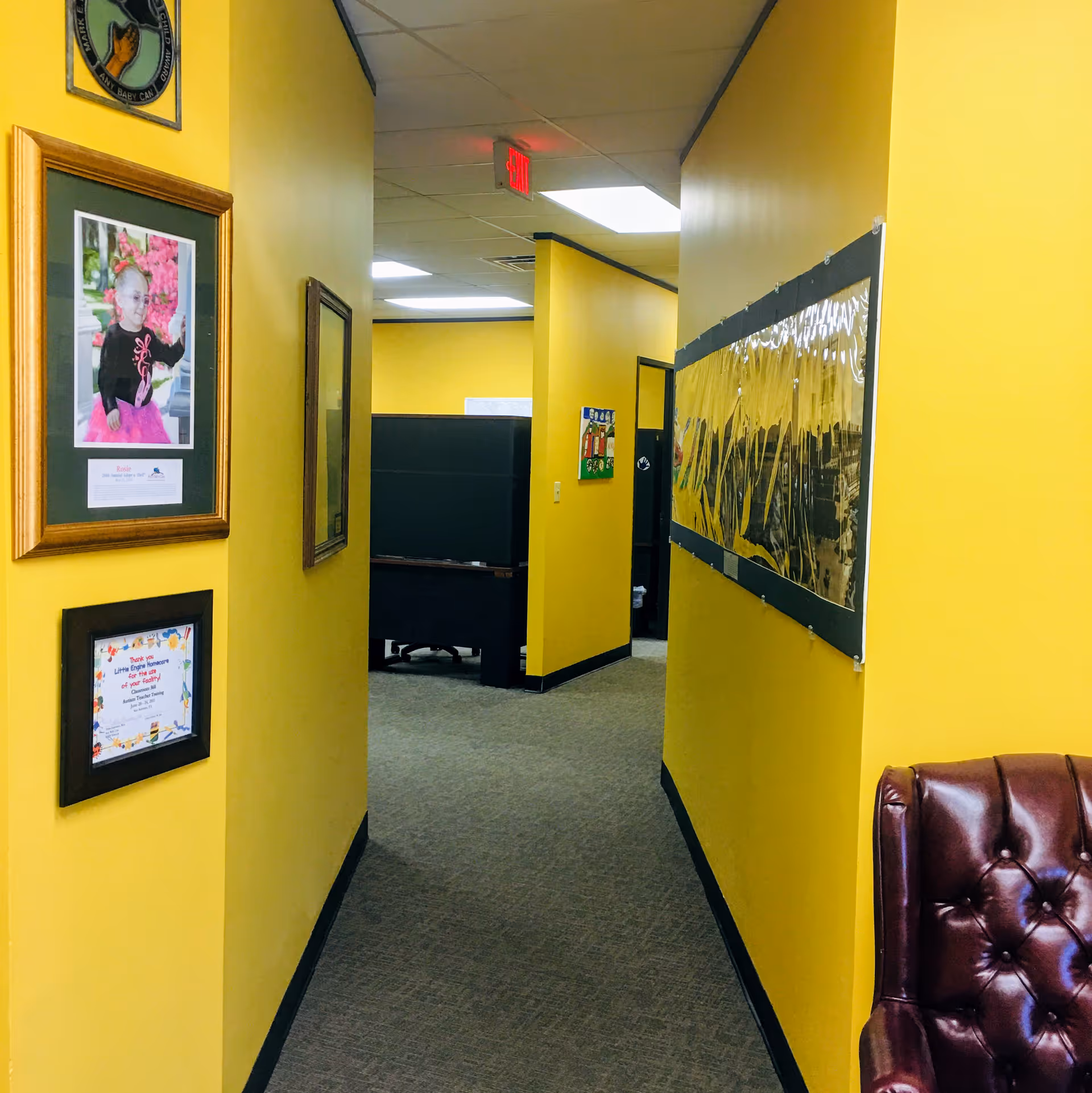 Yellow-walled hallway of a senior living facility with framed pictures and a brown chair.