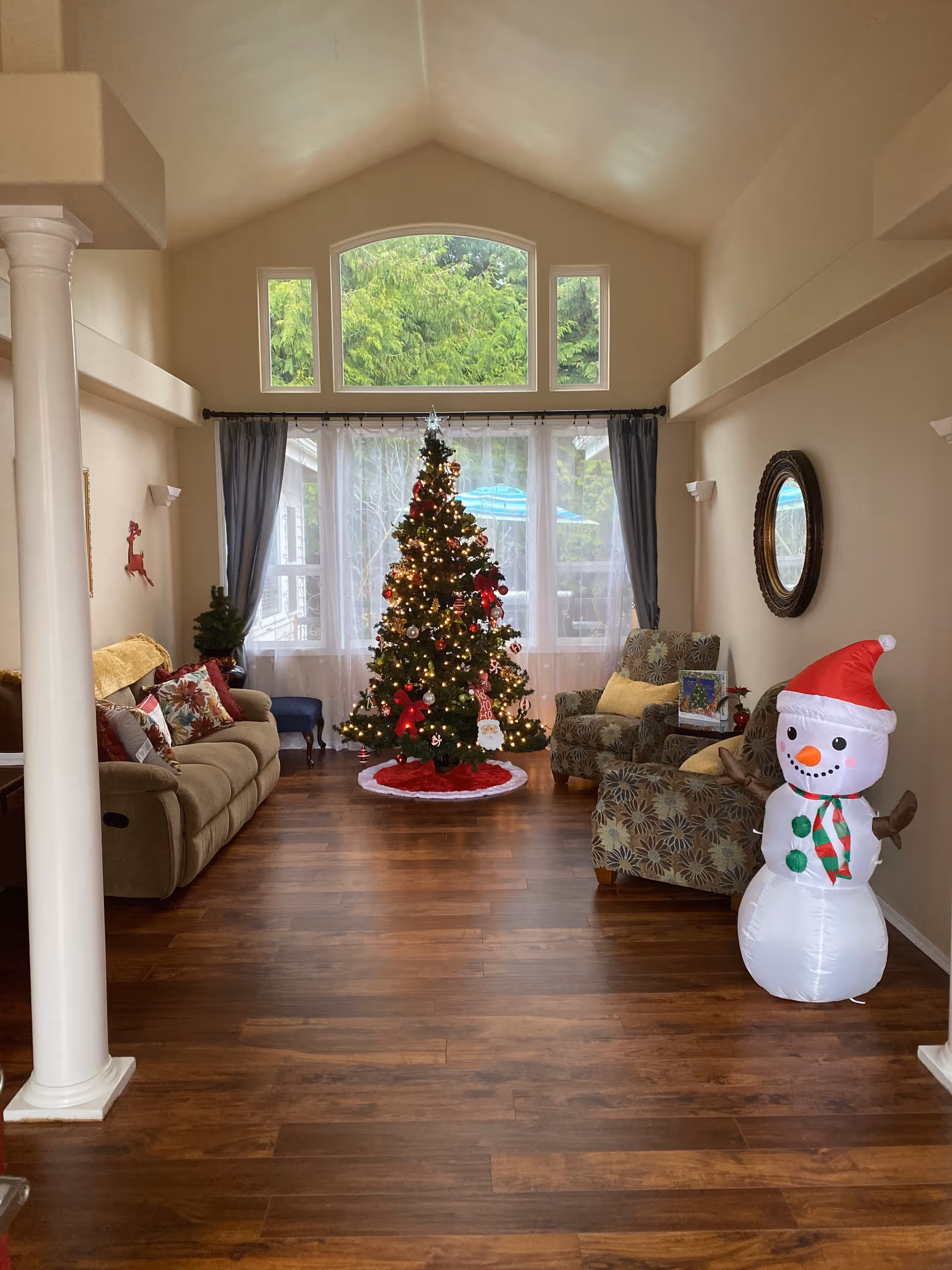 Festive living room with a decorated Christmas tree, snowman, couches, and large windows.
