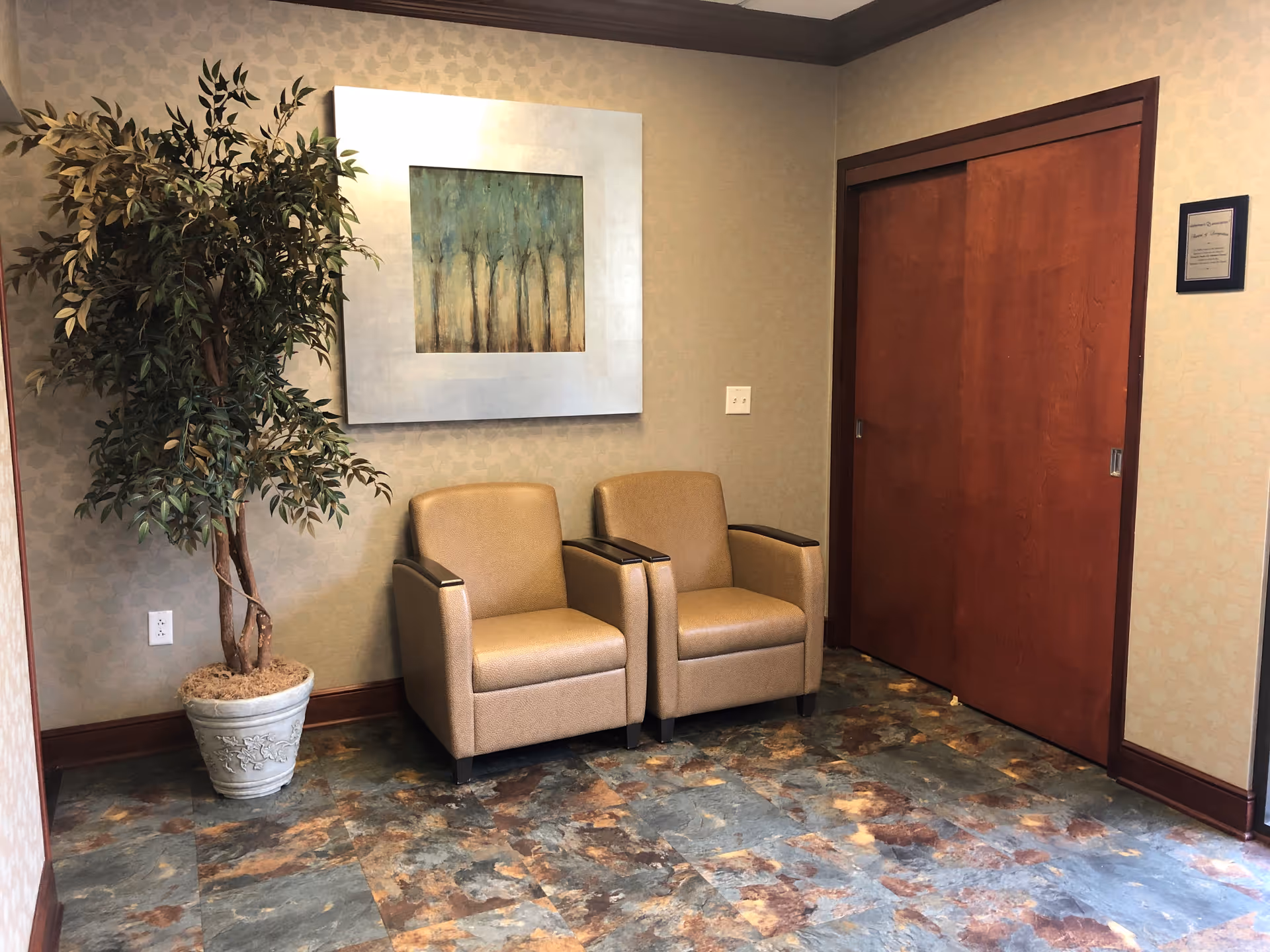 Two beige chairs sit beside a potted plant, framed art, and a wooden door.