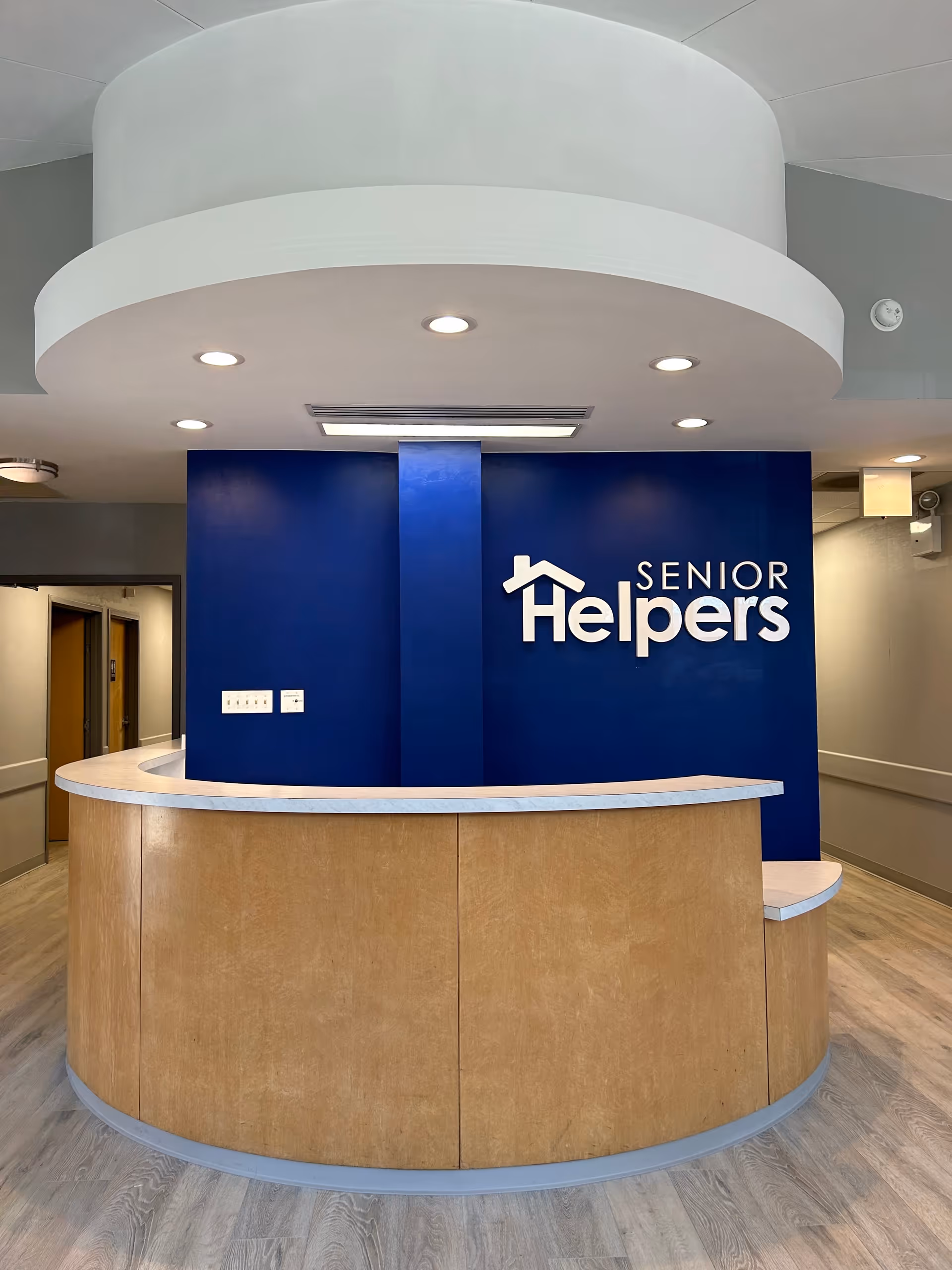 Curved wooden reception desk sits in a blue-walled interior lobby with a Senior Helpers sign.