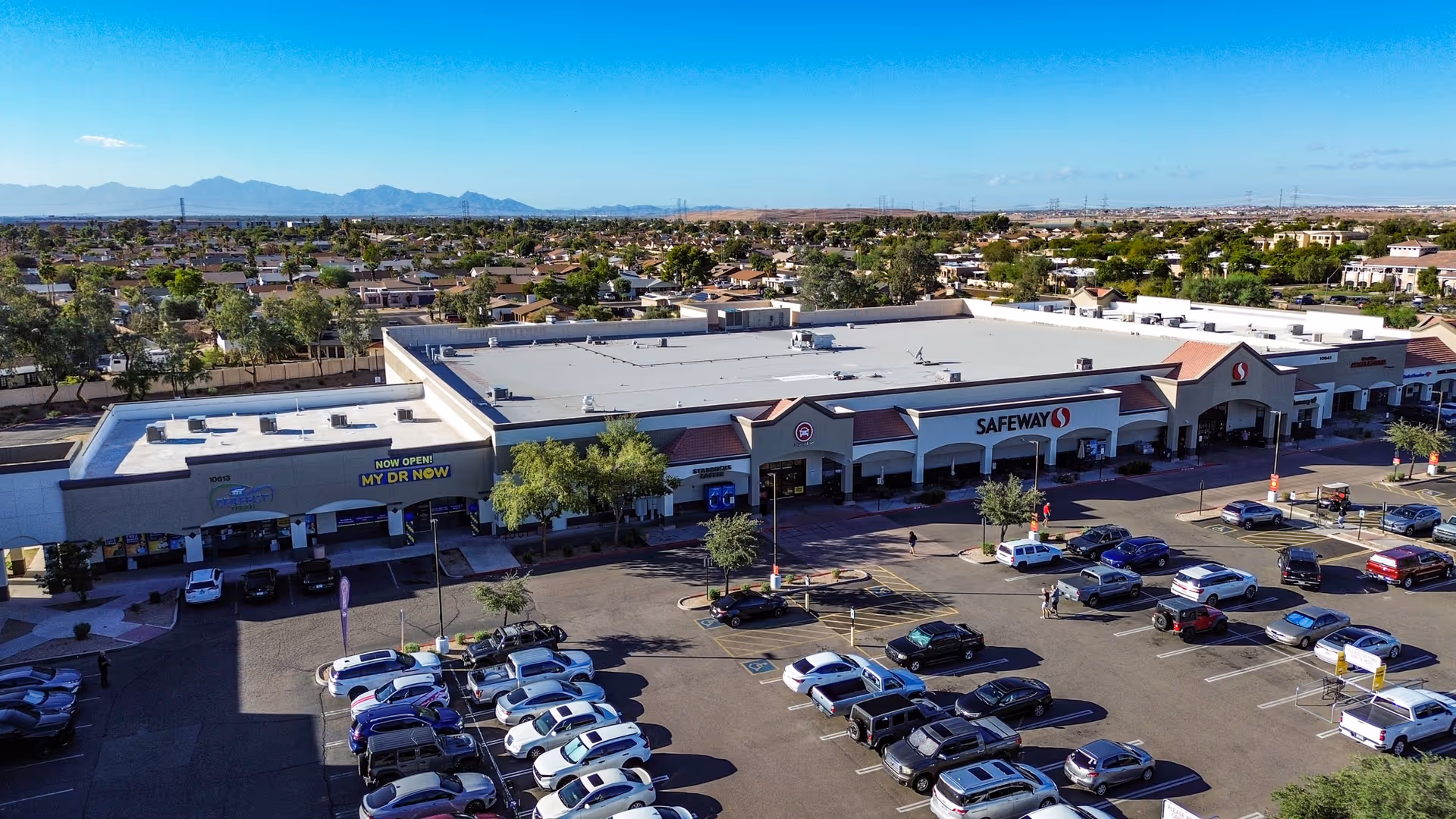 Exterior of a shopping center with MY DR NOW and Safeway storefronts, and numerous parked cars.