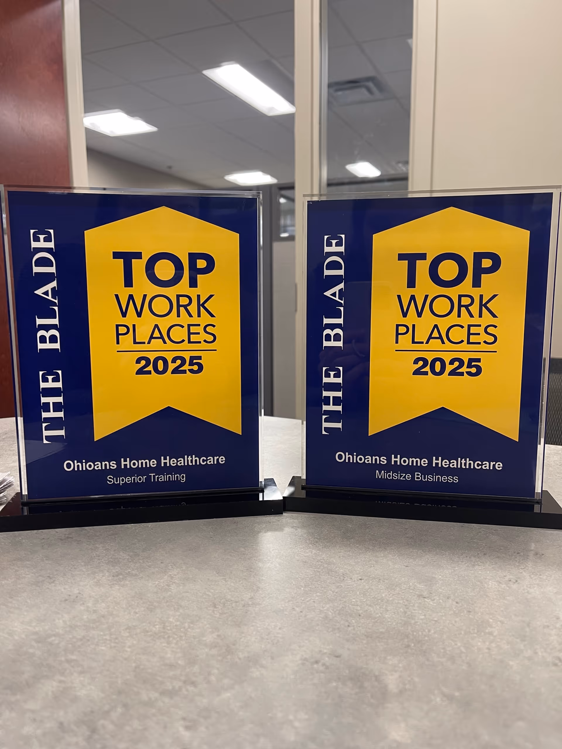 Two blue and yellow 'Top Work Places 2025' plaques on a desk in an office.