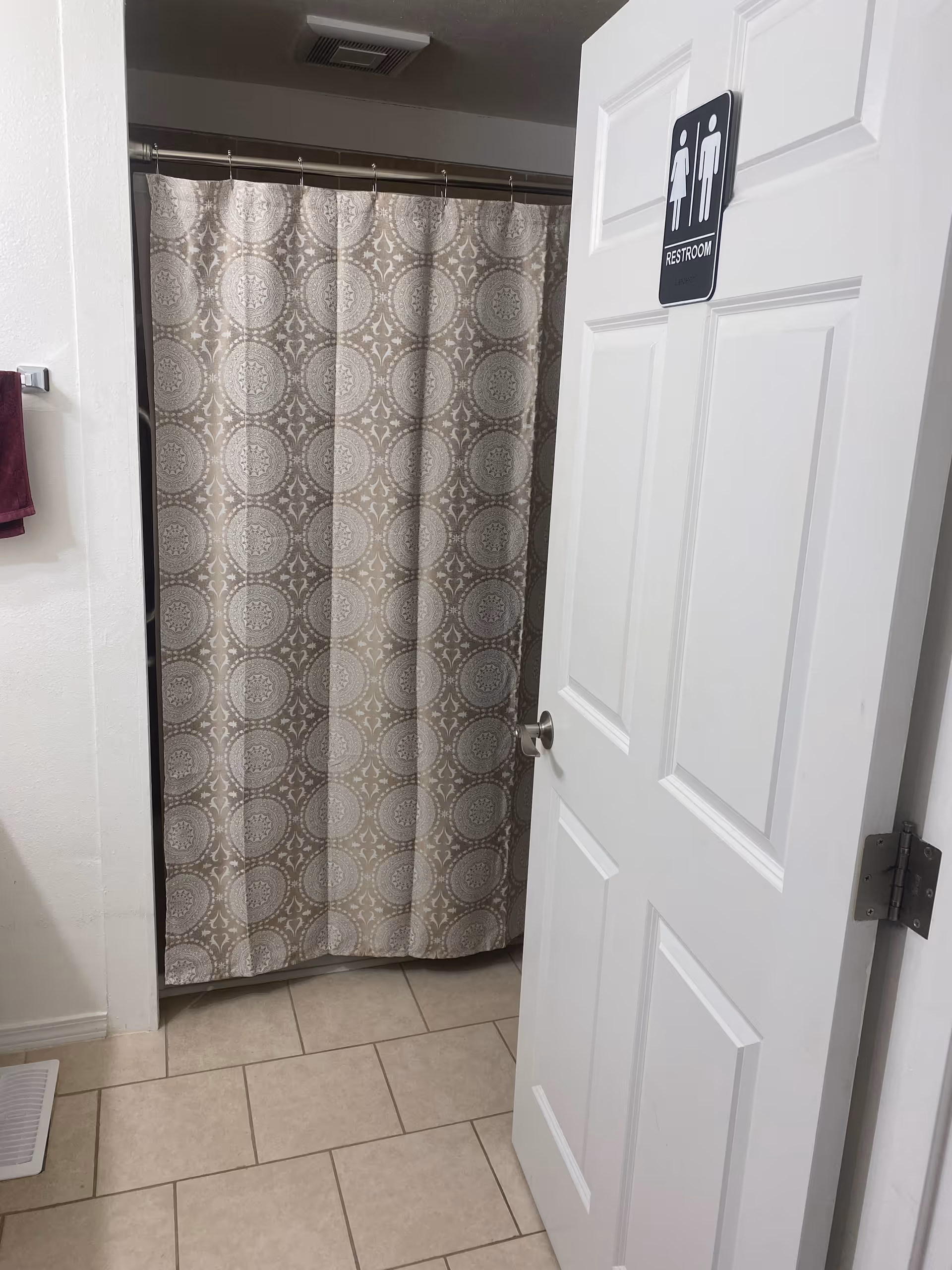 A bathroom door opens to a shower with a patterned curtain and tiled floor.