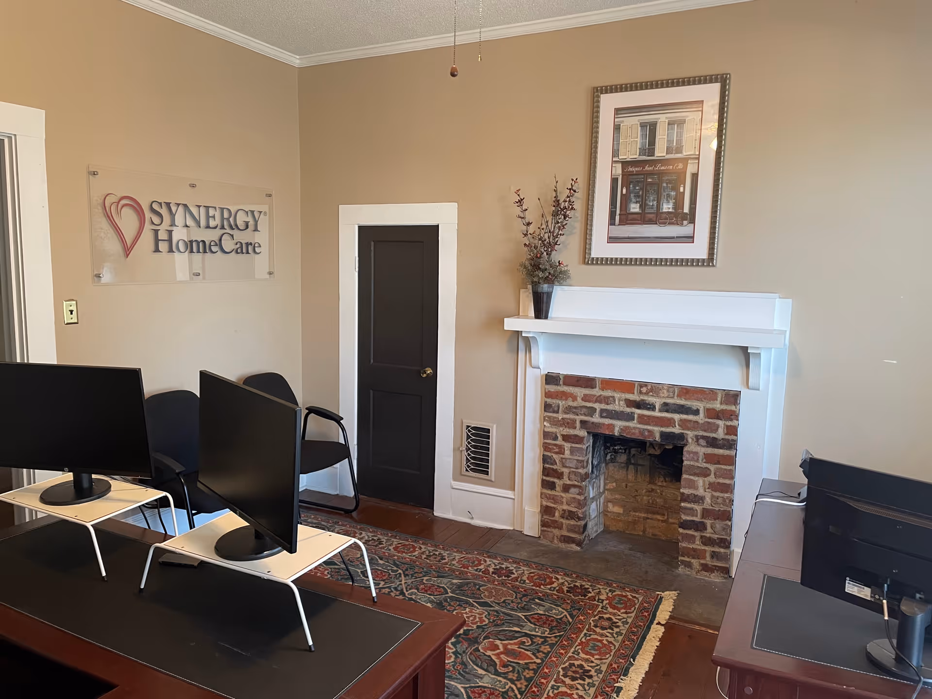 Office area with two computer desks, a fireplace, and a Synergy HomeCare sign on the wall.