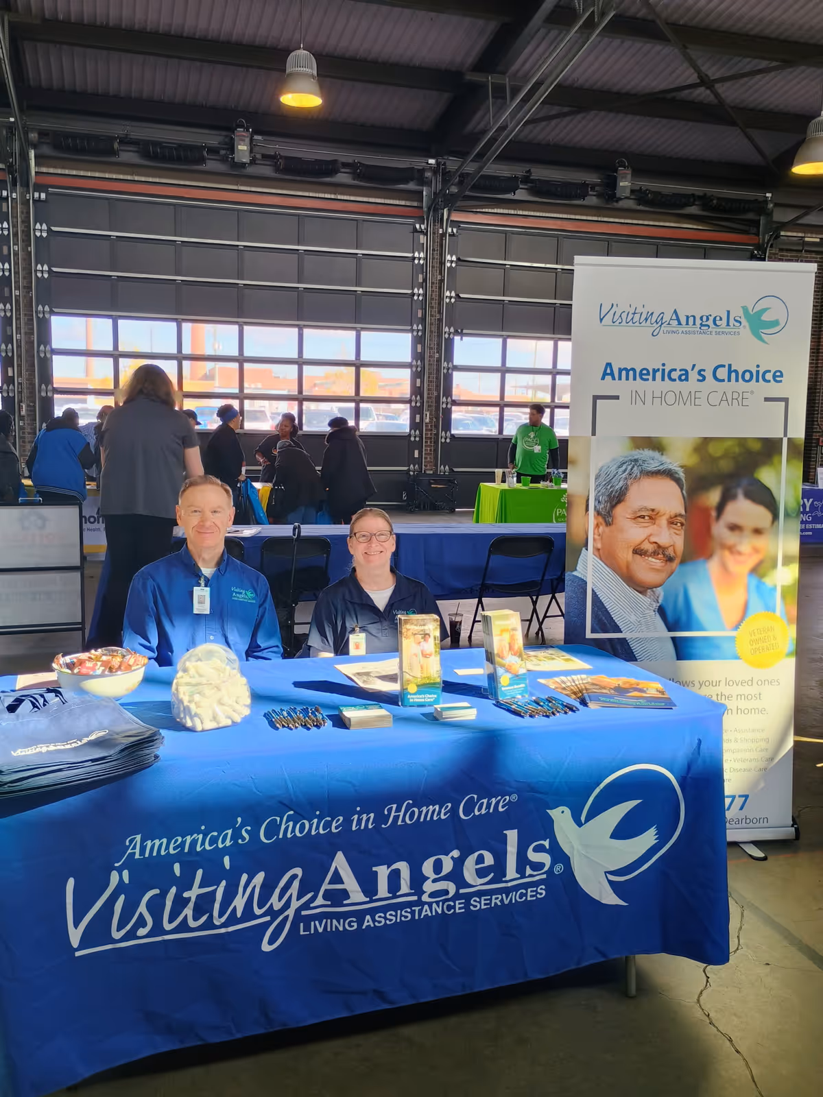 Promotional Visiting Angels booth with staff seated at a blue table inside an indoor event space.