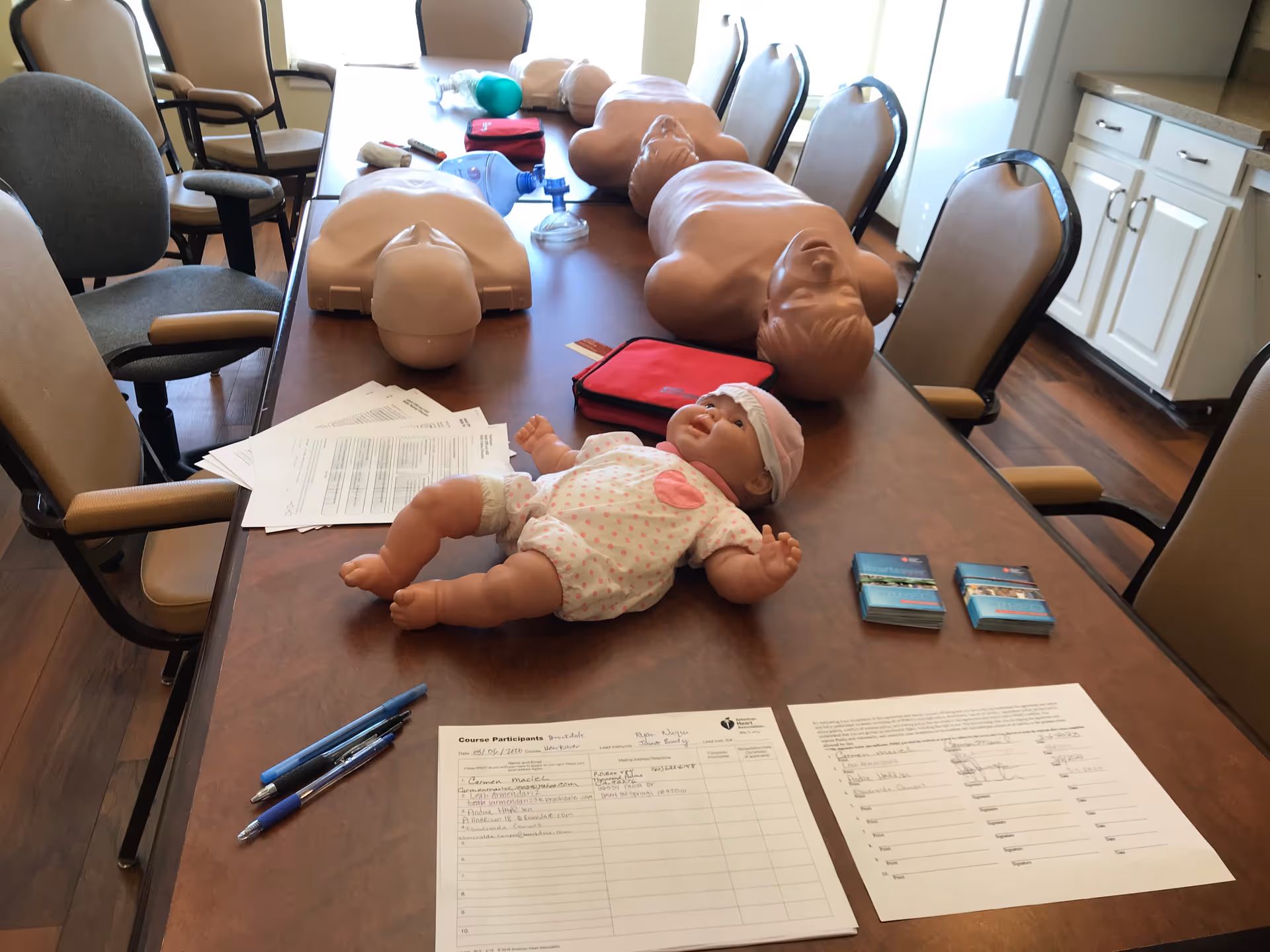 A CPR training room with a long table, mannequins, a baby doll, and papers.