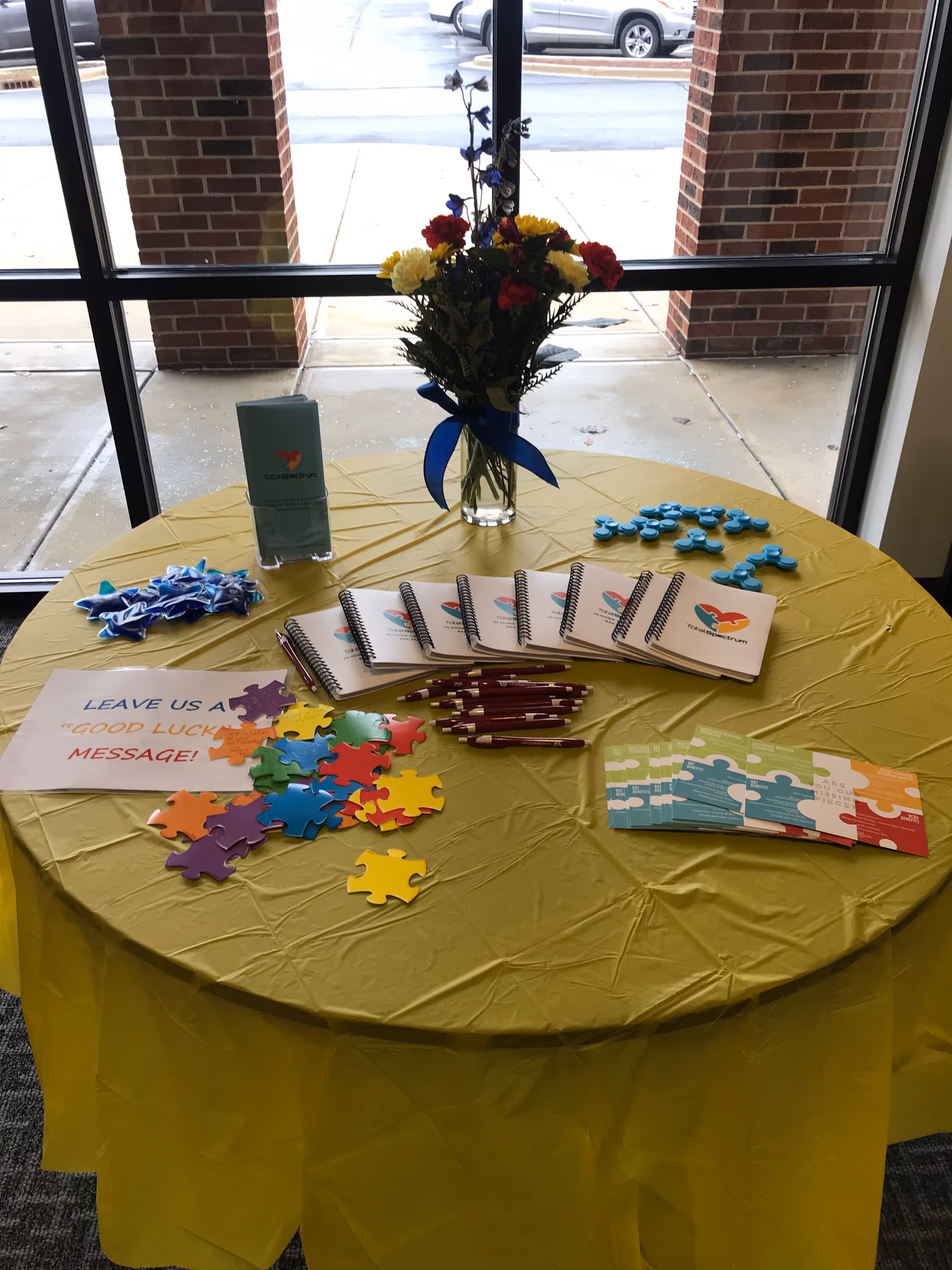 A round table with yellow cloth holds puzzles, notebooks, pens, and a bouquet near a window.