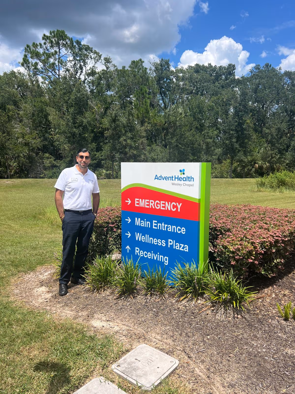 A man stands beside a colorful AdventHealth sign on a grassy outdoor area.