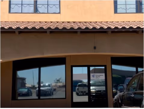 Front entrance of a tan stucco building with windows and a tiled roof, cars parked outside.