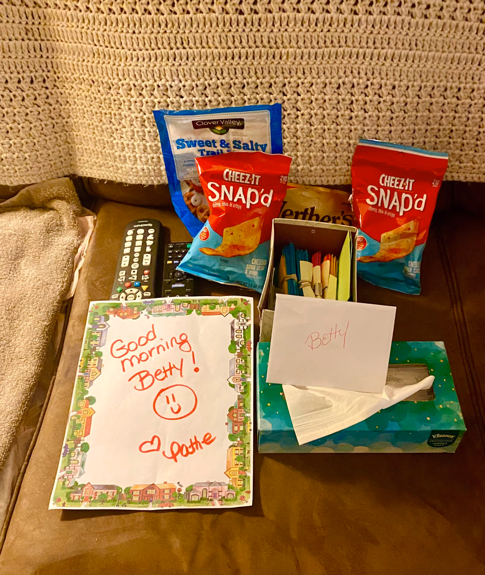 Living room couch with snacks, a tissue box, remote, and a 'Good morning Betty!' note.