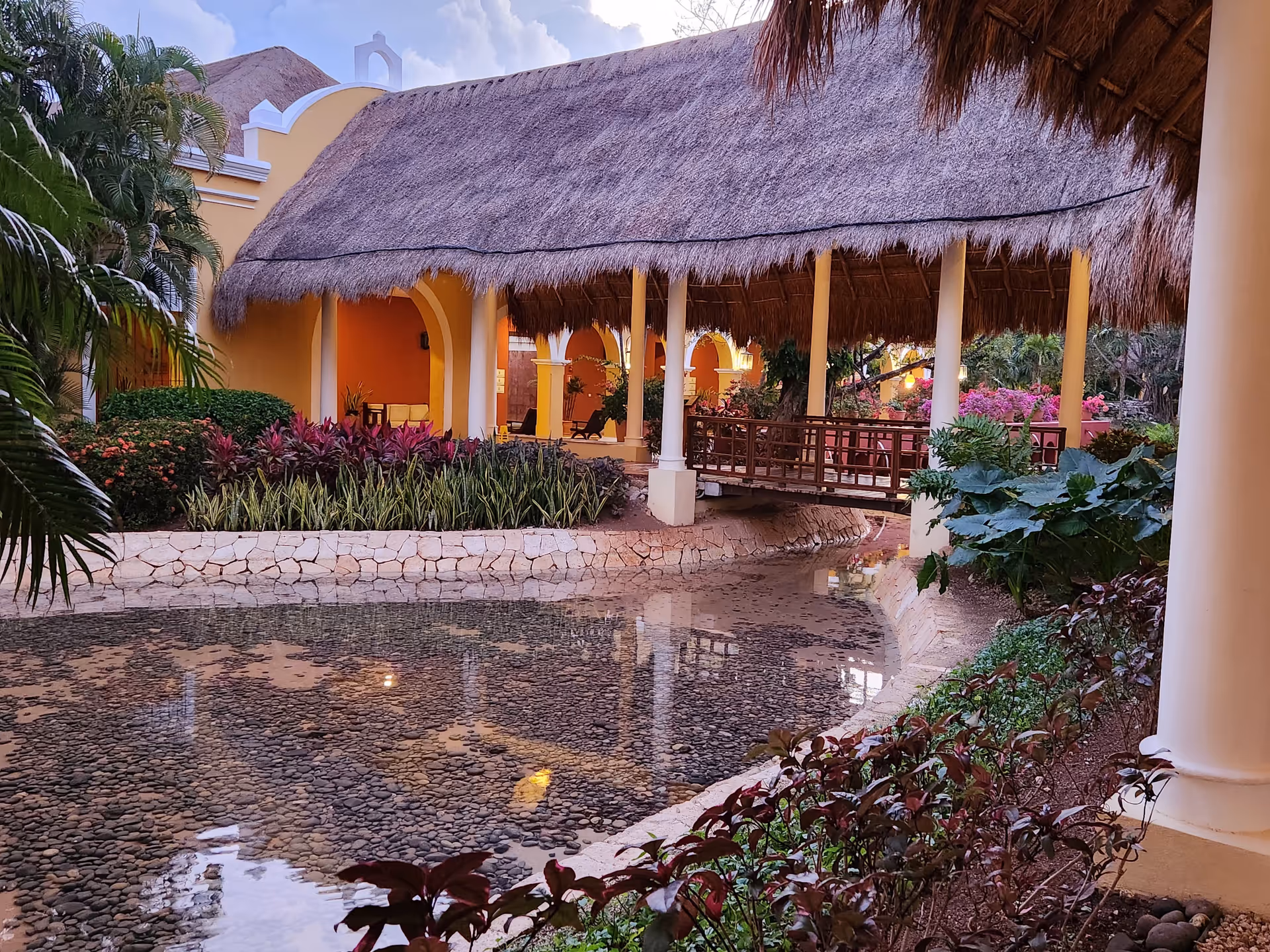 Yellow building exterior with thatched pavilion, water feature, and lush landscaping.