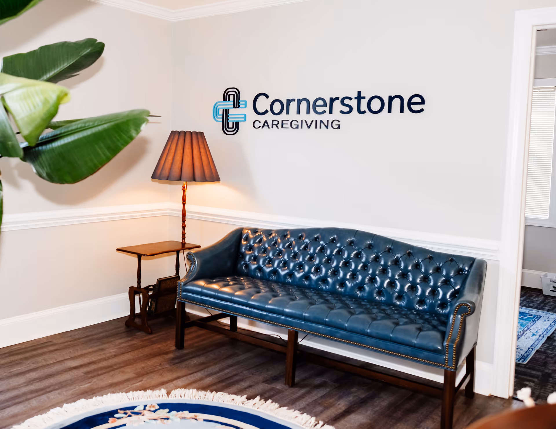 Cornerstone Caregiving lobby with a blue tufted sofa, lamp, plant, and wall logo.