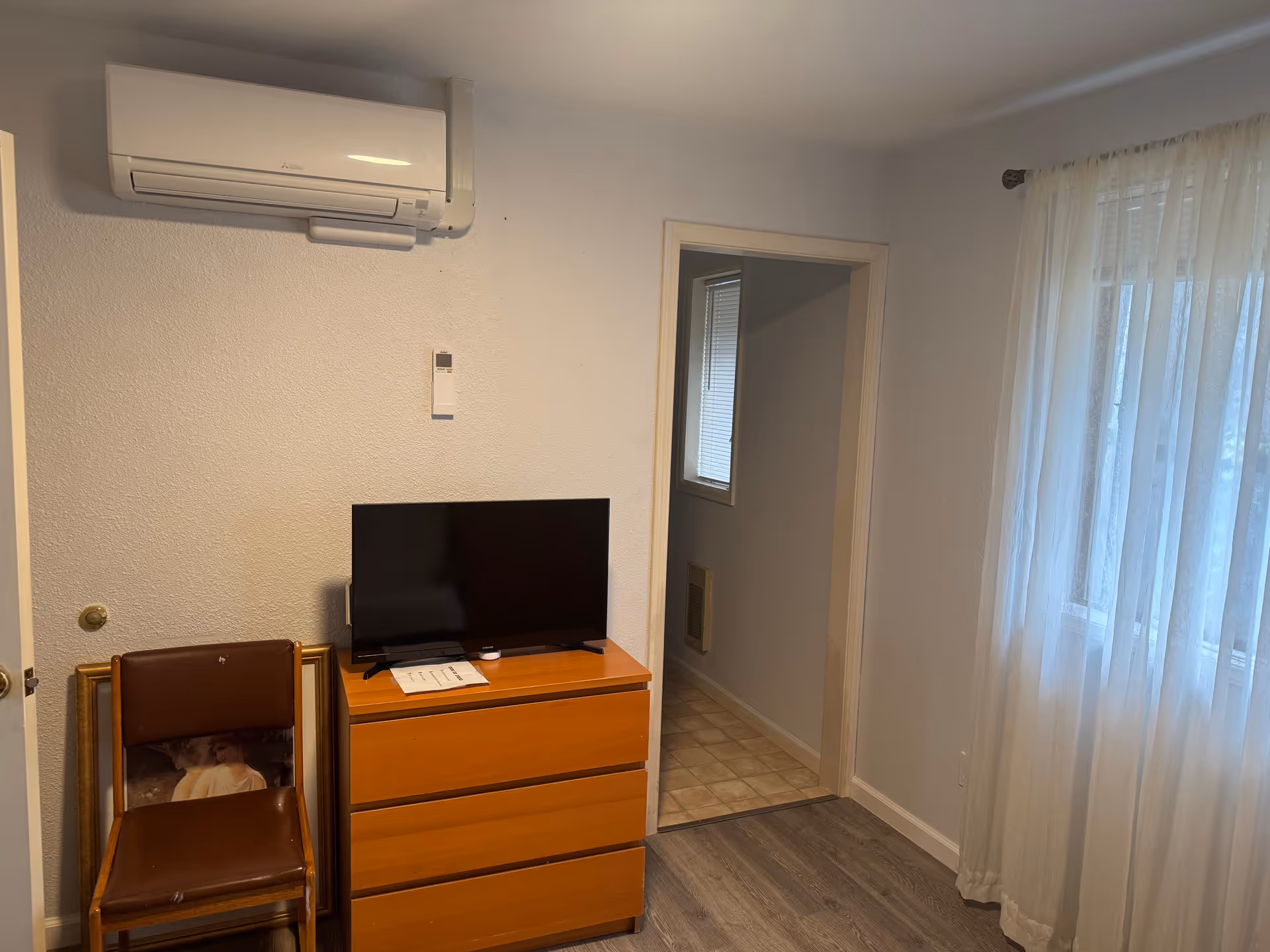 A small furnished room with a dresser, TV, chair, air conditioner, and sheer curtains.