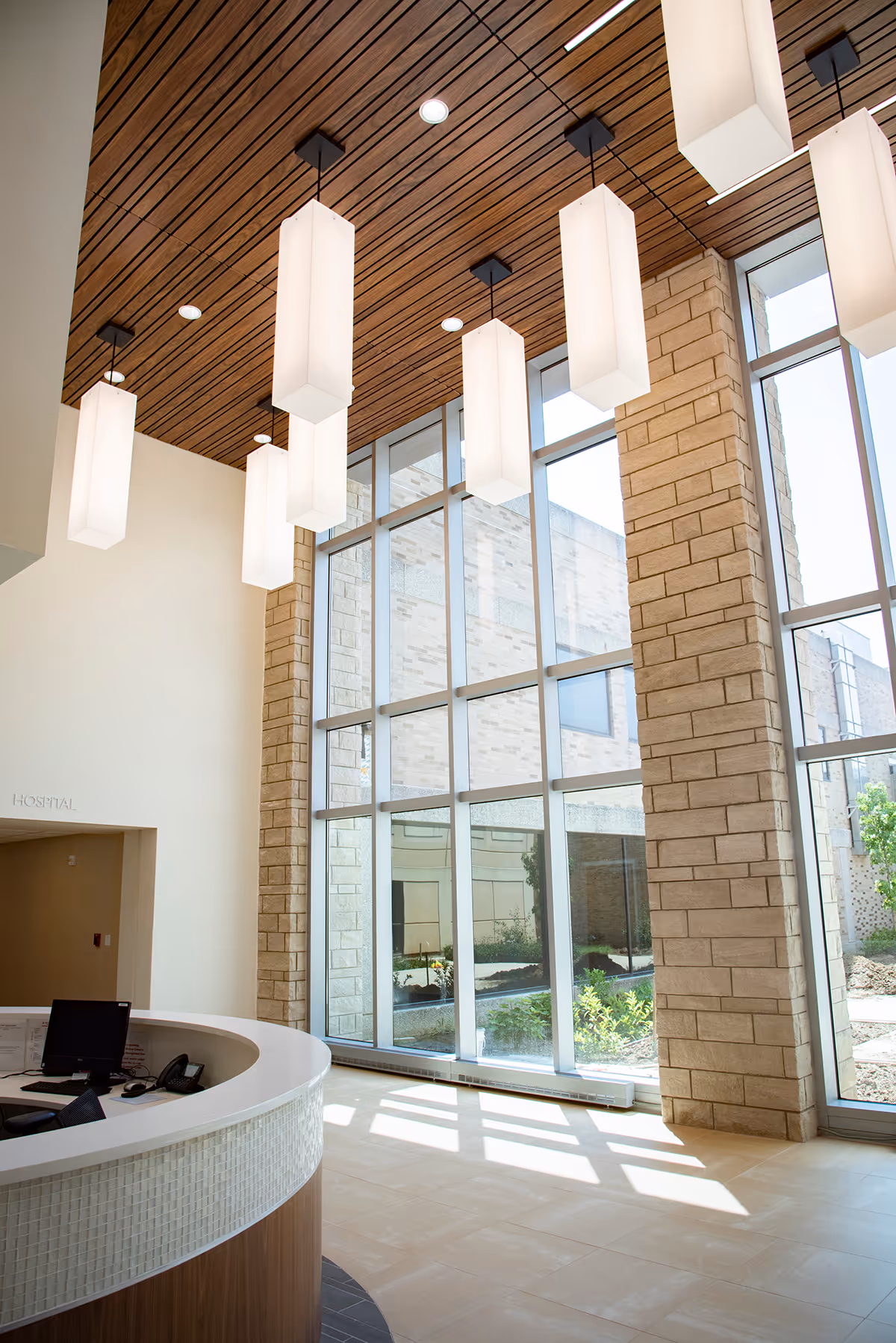 Modern hospital reception lobby with tall windows, brick columns, and hanging pendant lights.