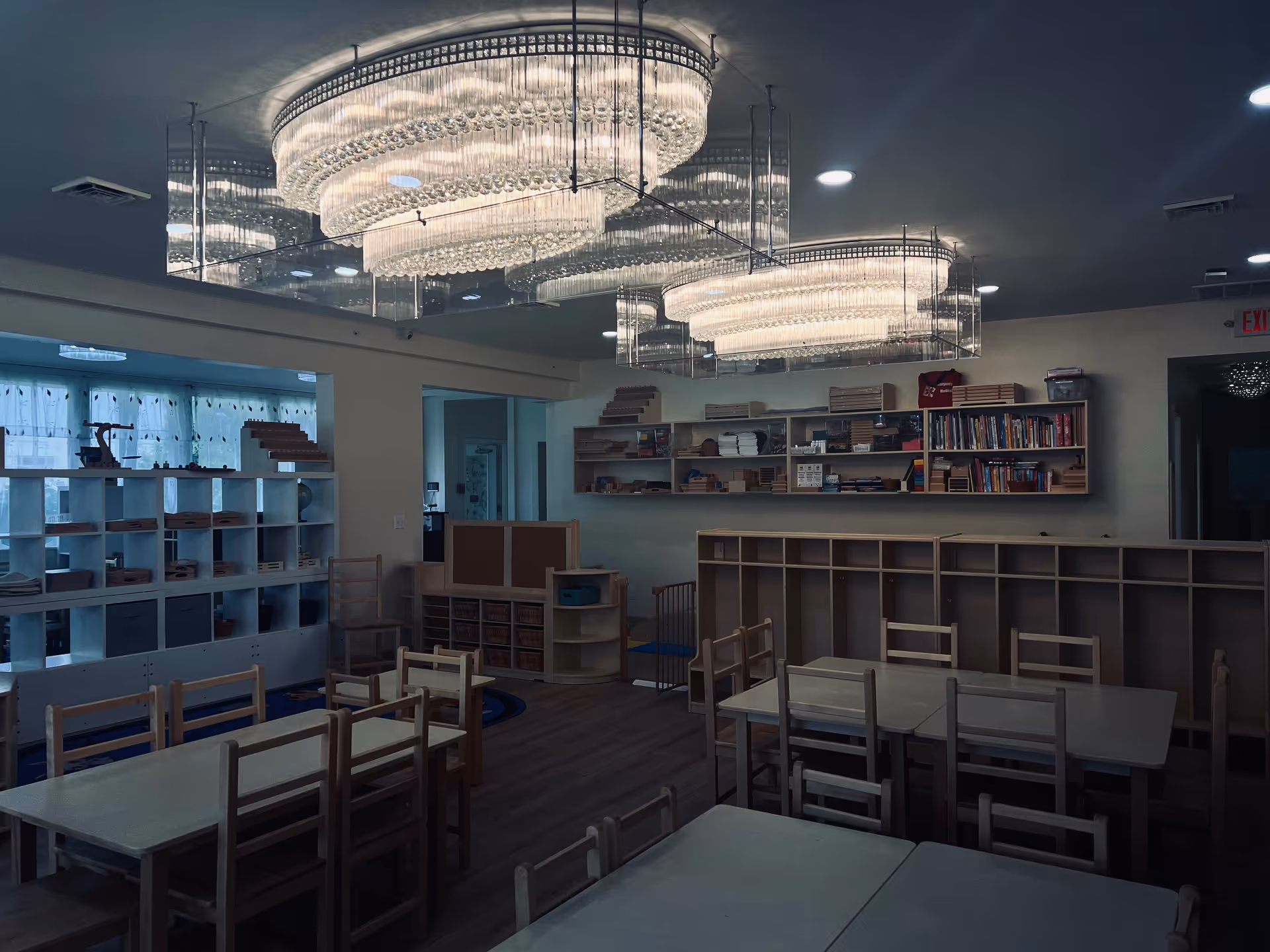 Interior of a day center with small tables, wooden chairs, shelves, and chandeliers.