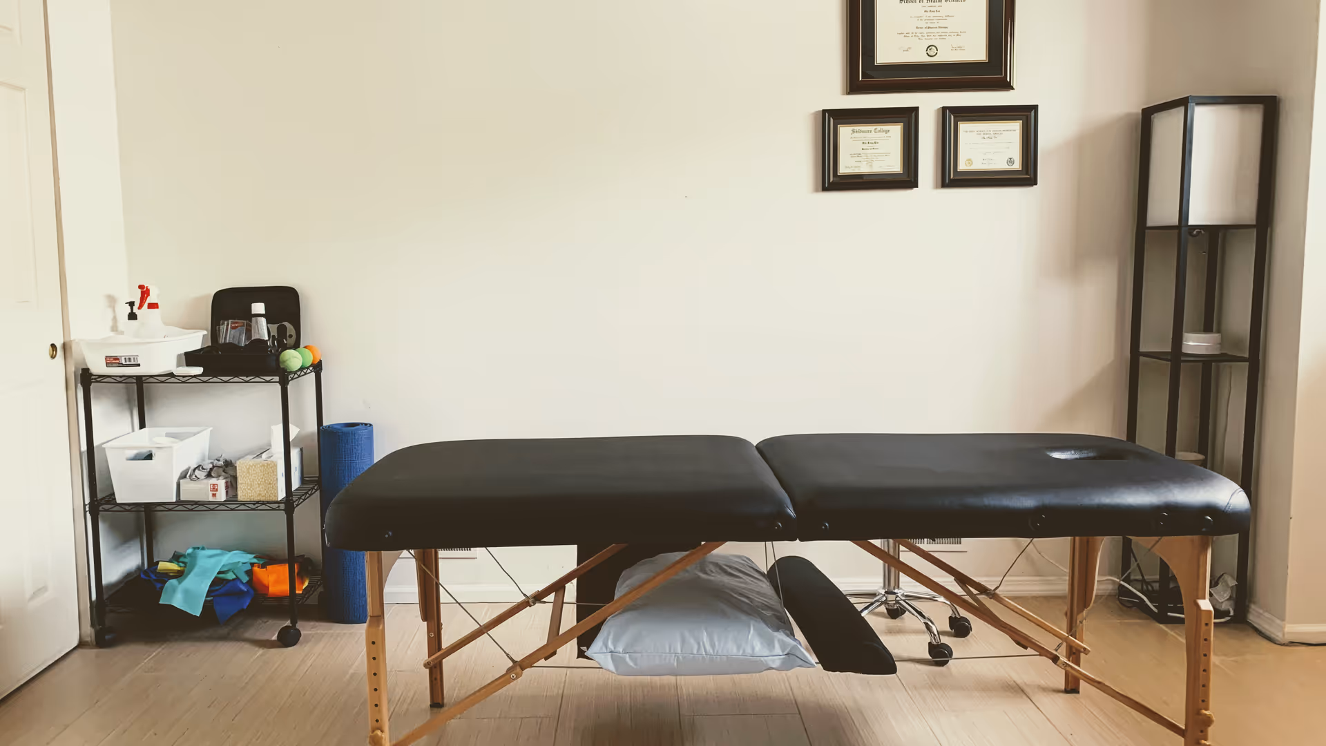 A black therapy table in a tidy room with shelves and framed certificates.