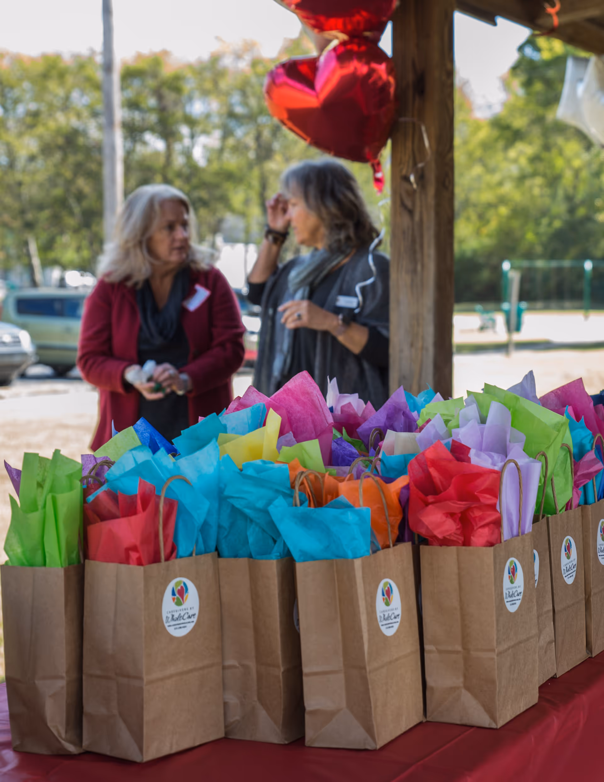Colorful gift bags lined up on a table outdoors under a wooden pavilion.